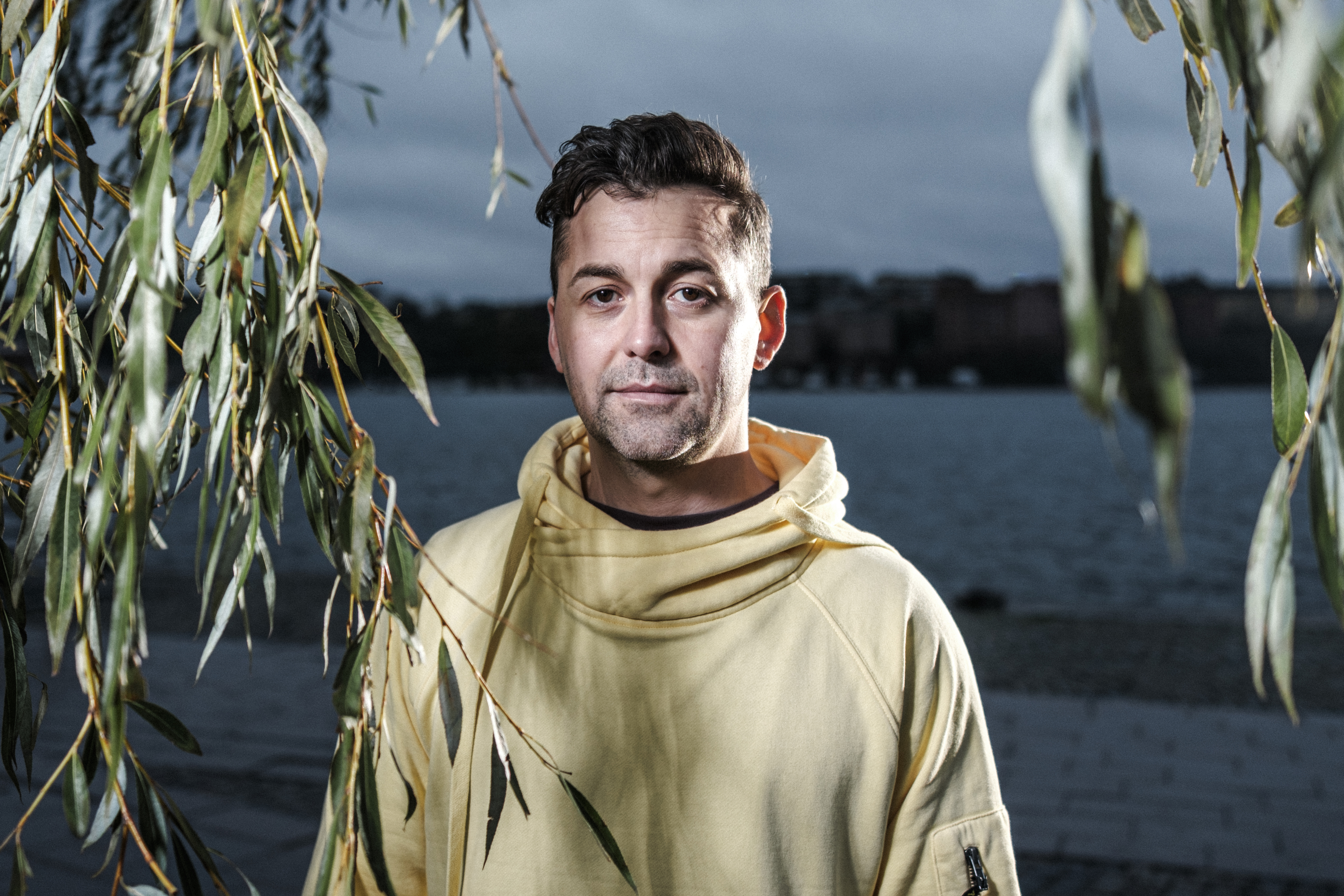 Joacim Karlsson i gul huvtröja står nära vatten, delvis skymd av blad, med molnig himmel i bakgrunden.