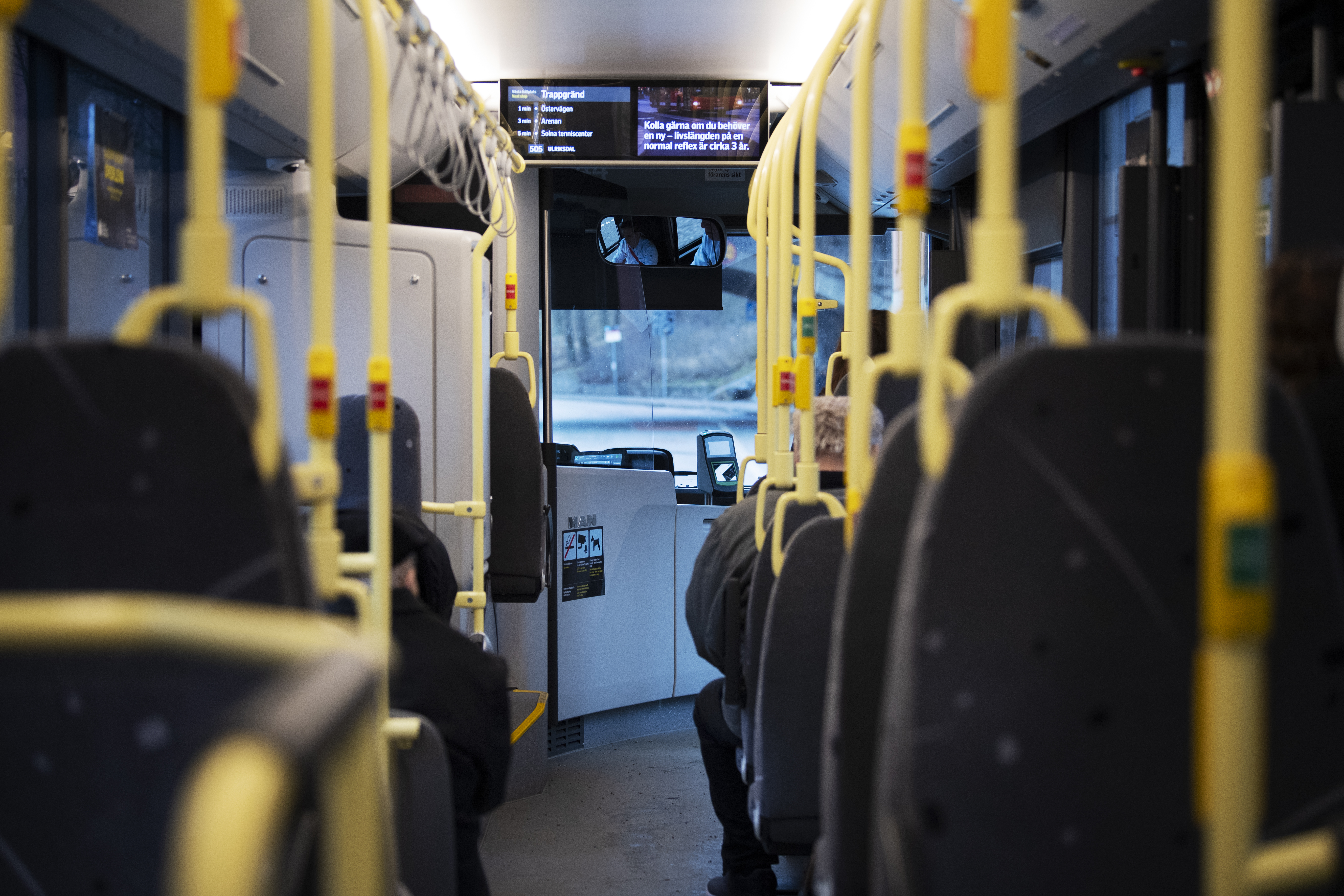 Insidan av en stadsbuss med gula handtag, några sittande passagerare och en digital skylt som visar ruttinformation längst fram.