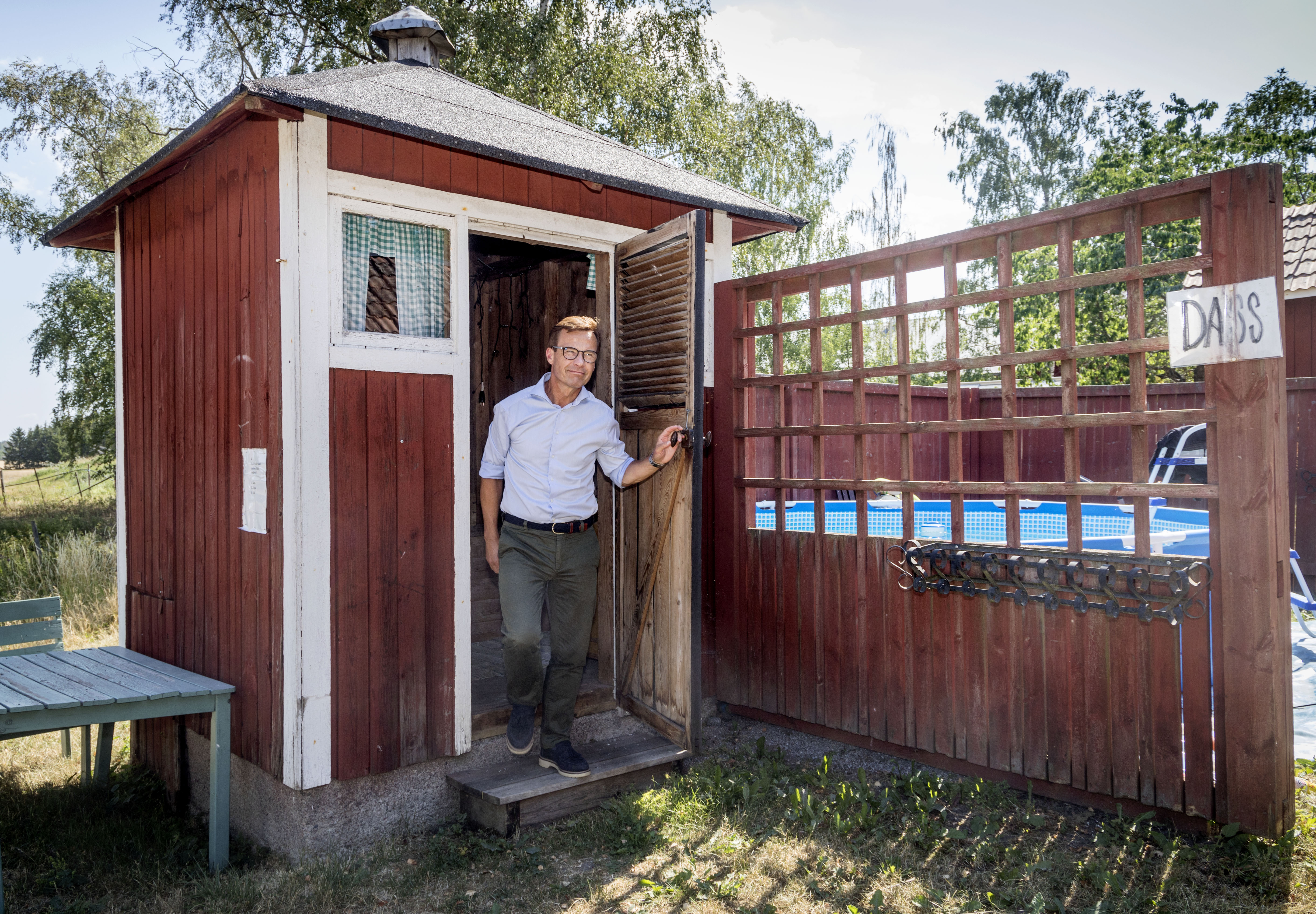 En man med glasögon och skjorta går ut ur ett rött utedass märkt "DASS" vid ett staket, med en pool i bakgrunden.