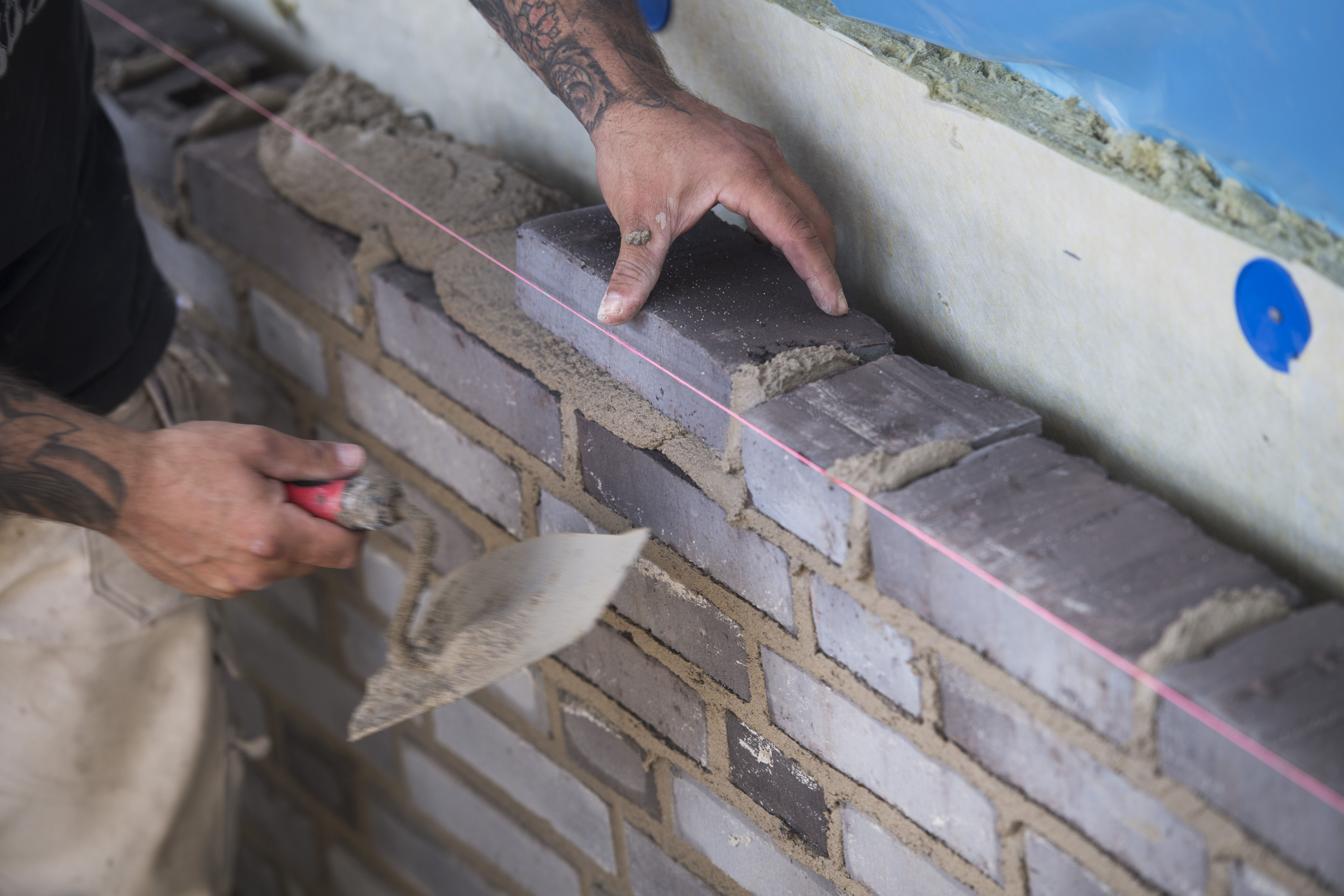 En person använder en murslev för att lägga murbruk och stapla tegel vid byggandet av en tegelvägg, med ett snöre för riktning.