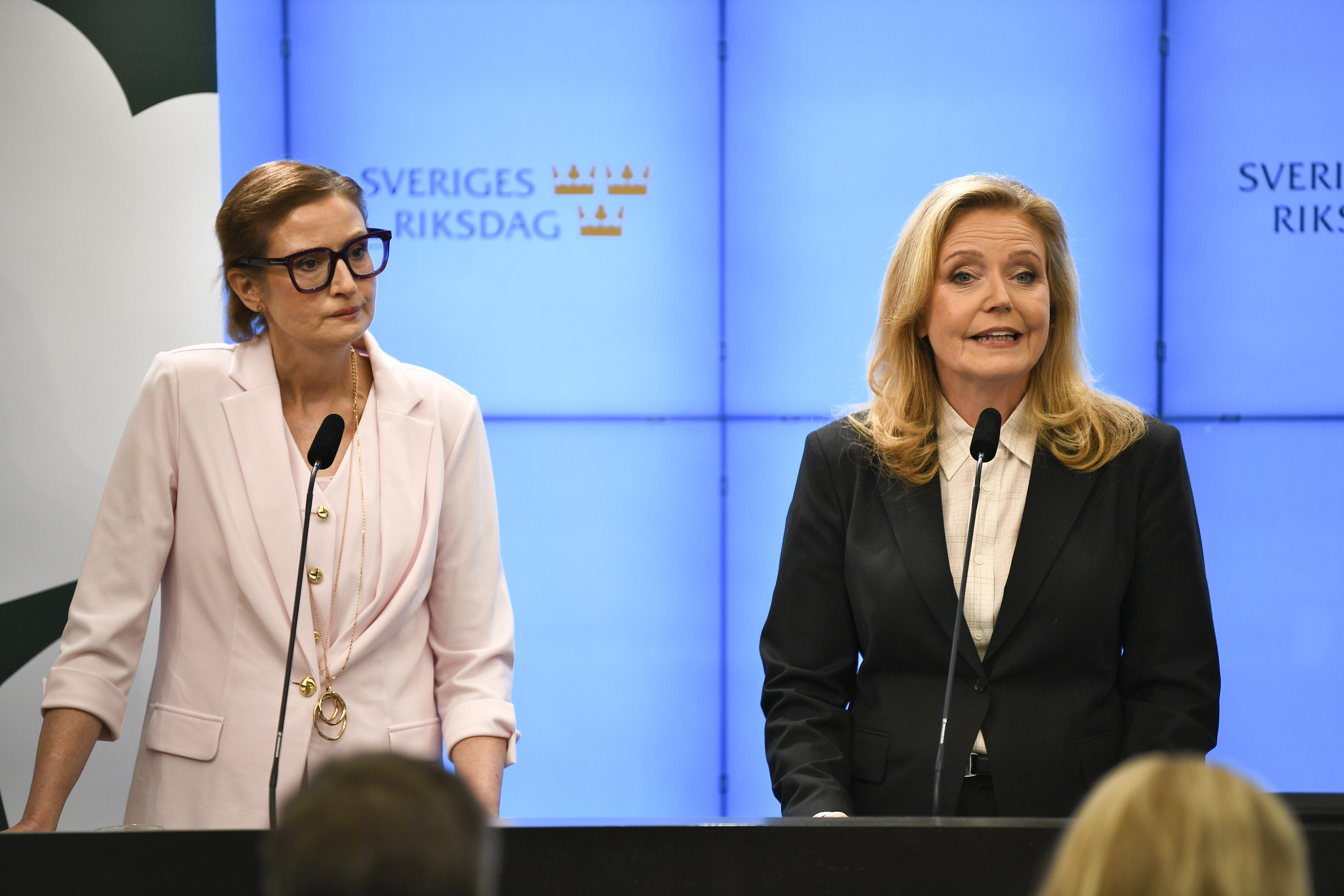 Två kvinnor står vid talarpodium och talar vid en presskonferens framför skärmar med Sveriges riksdags logotyp.