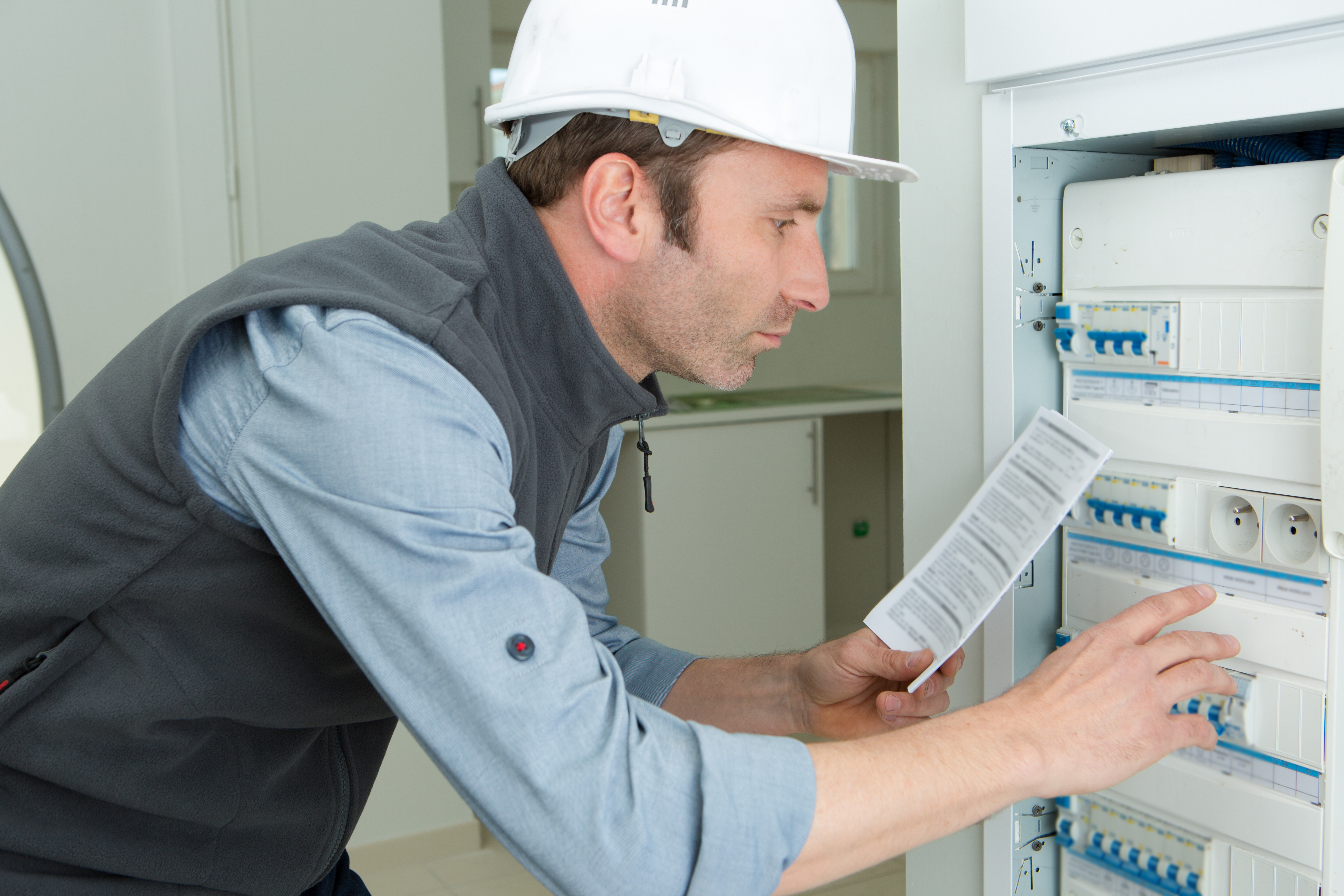 En elektriker med vit hjälm och grå väst granskar en elcentral och håller i ett papper med instruktioner.
