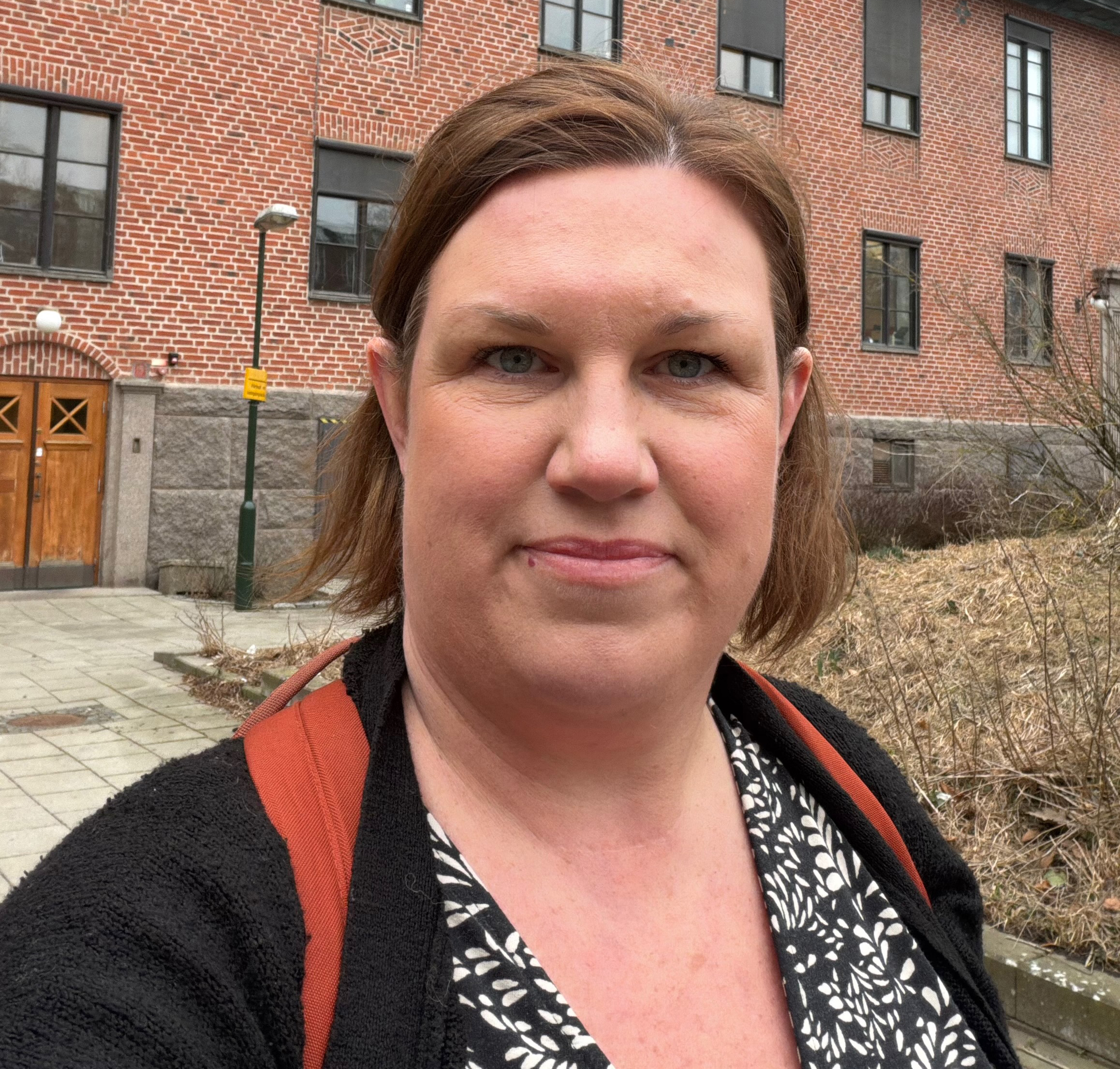En person med kort brunt hår och mönstrad tröja står utomhus framför en tegelbyggnad och tittar mot kameran.