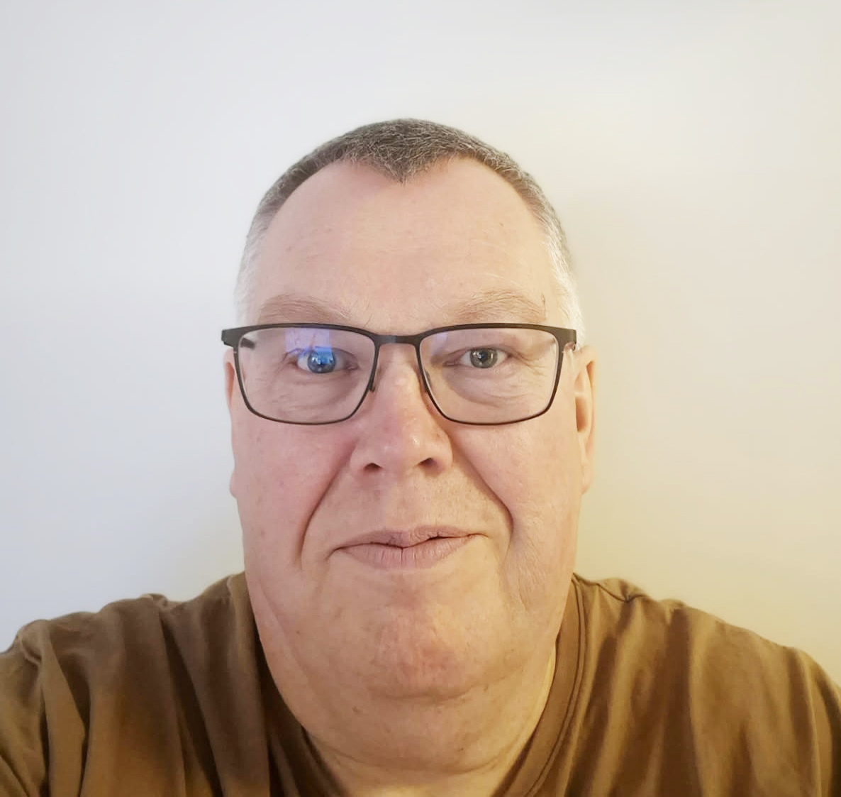 En medelålders man med kort grått hår och glasögon tittar mot kameran, har på sig en brun skjorta mot ljus bakgrund.