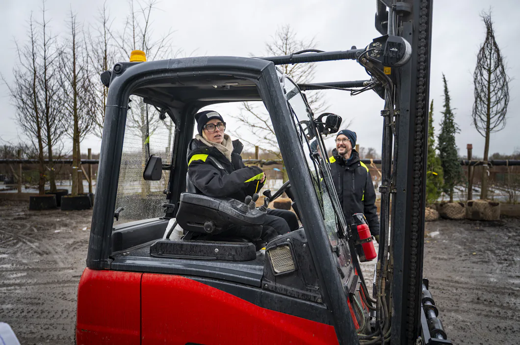 En person sitter i förarsätet på en röd gaffeltruck och den andra står bredvid gaffeltrucken utomhus med träd och växter i bakgrunden.