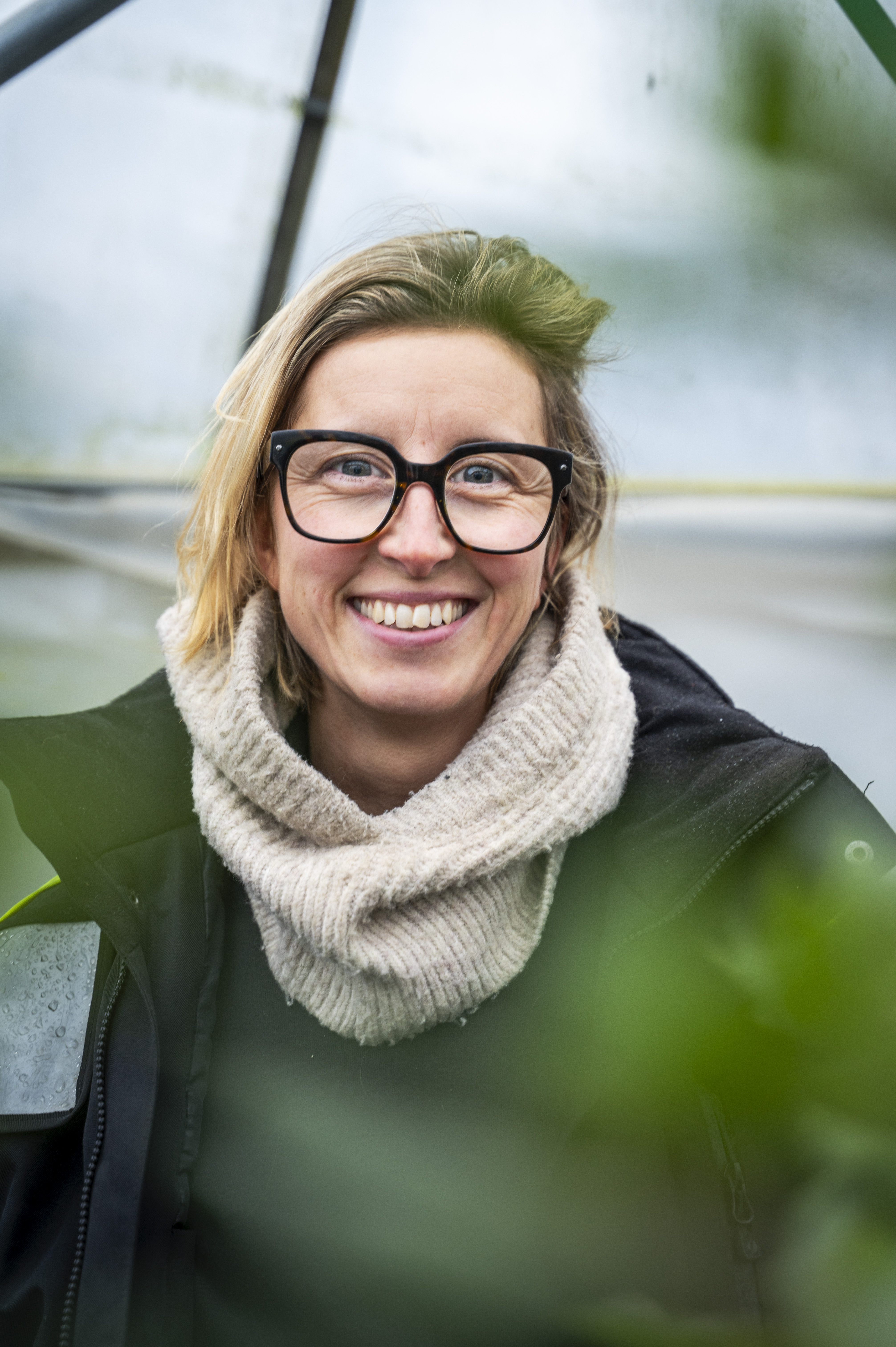 En kvinna med glasögon och en ljus scarf ler mot kameran och står utomhus med växter i förgrunden.