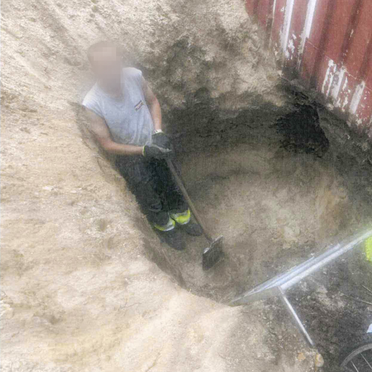 En person med maskat ansikte står i en grop i marken och håller en spade, bredvid en husvägg.