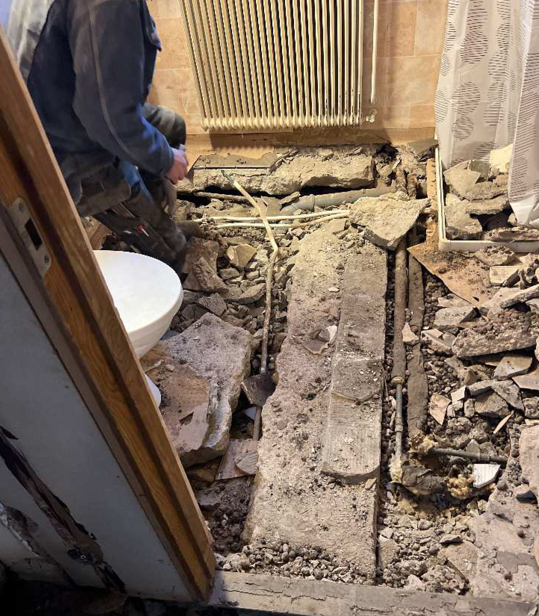 En person står i ett badrum där golvet är delvis borttaget, rör och skräp syns runt en toalett och ett element.