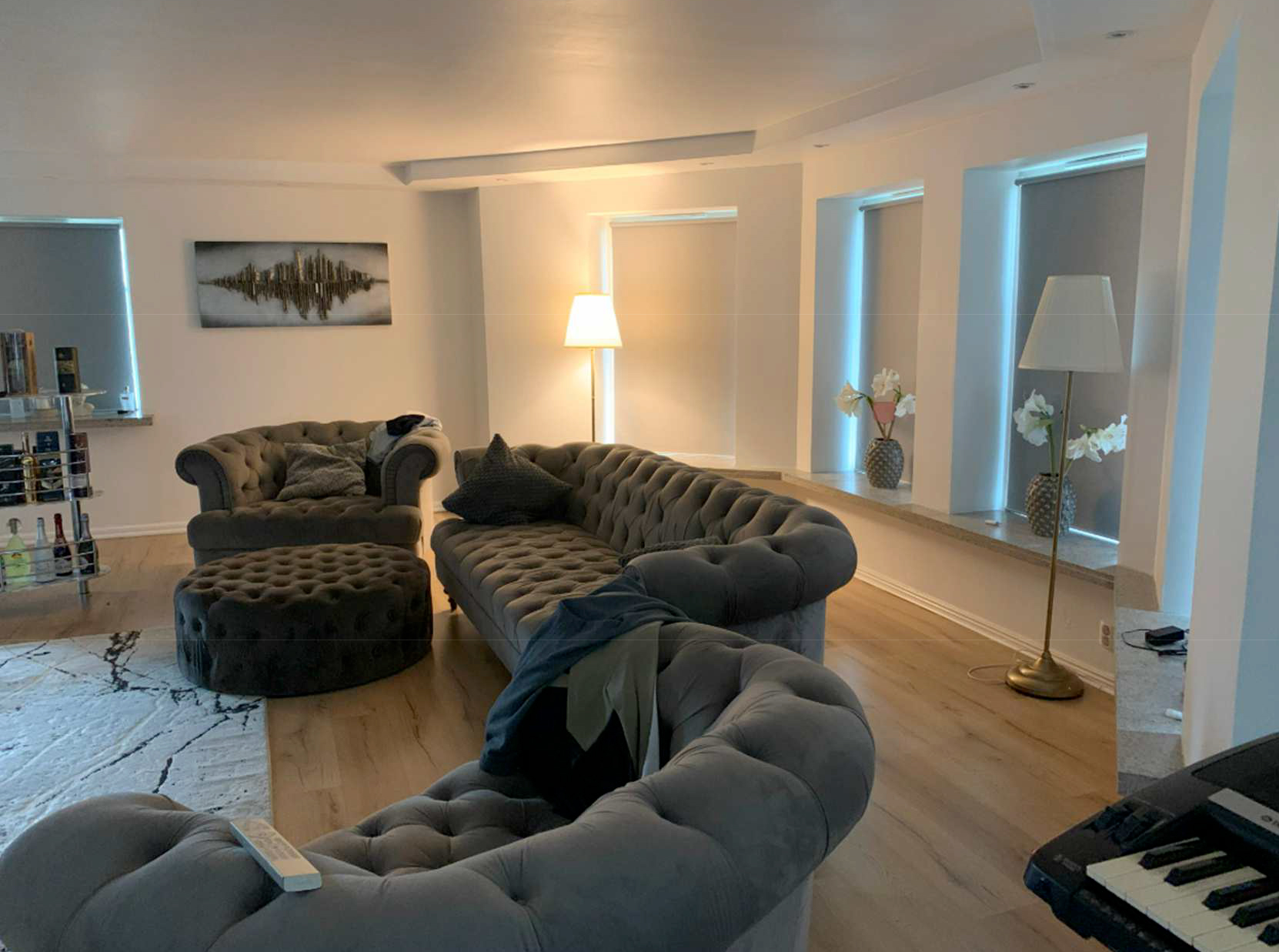 Vardagsrum med grå soffor, runt ottoman, två golvlampor, krukväxter med vita blommor på fönsterbrädet, tavla och keyboard i hörnet.