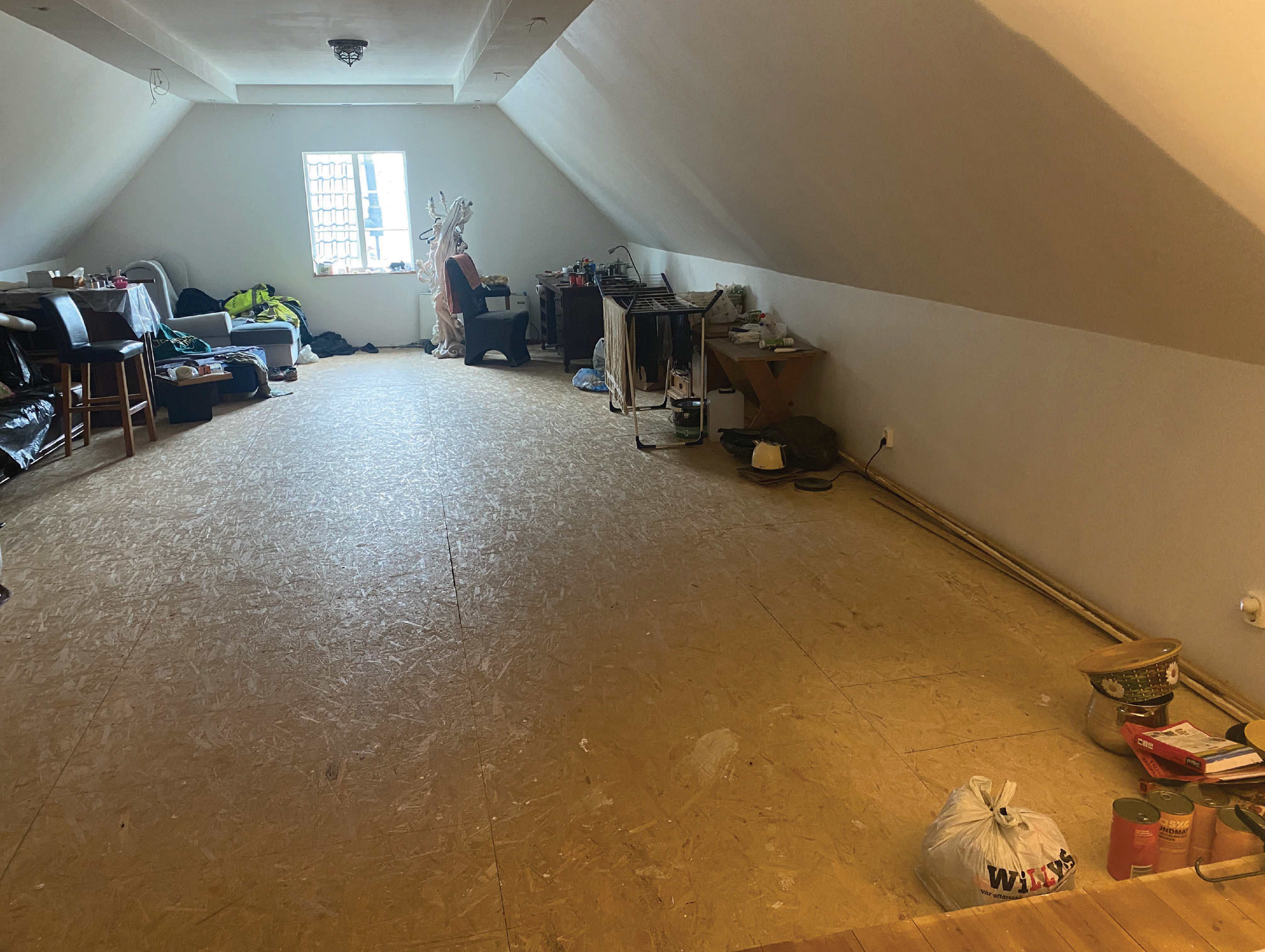 Ett vindsrum med ofärdigt golv, möbler utspridda och föremål längs väggarna. Ljus kommer in genom ett fönster i änden.