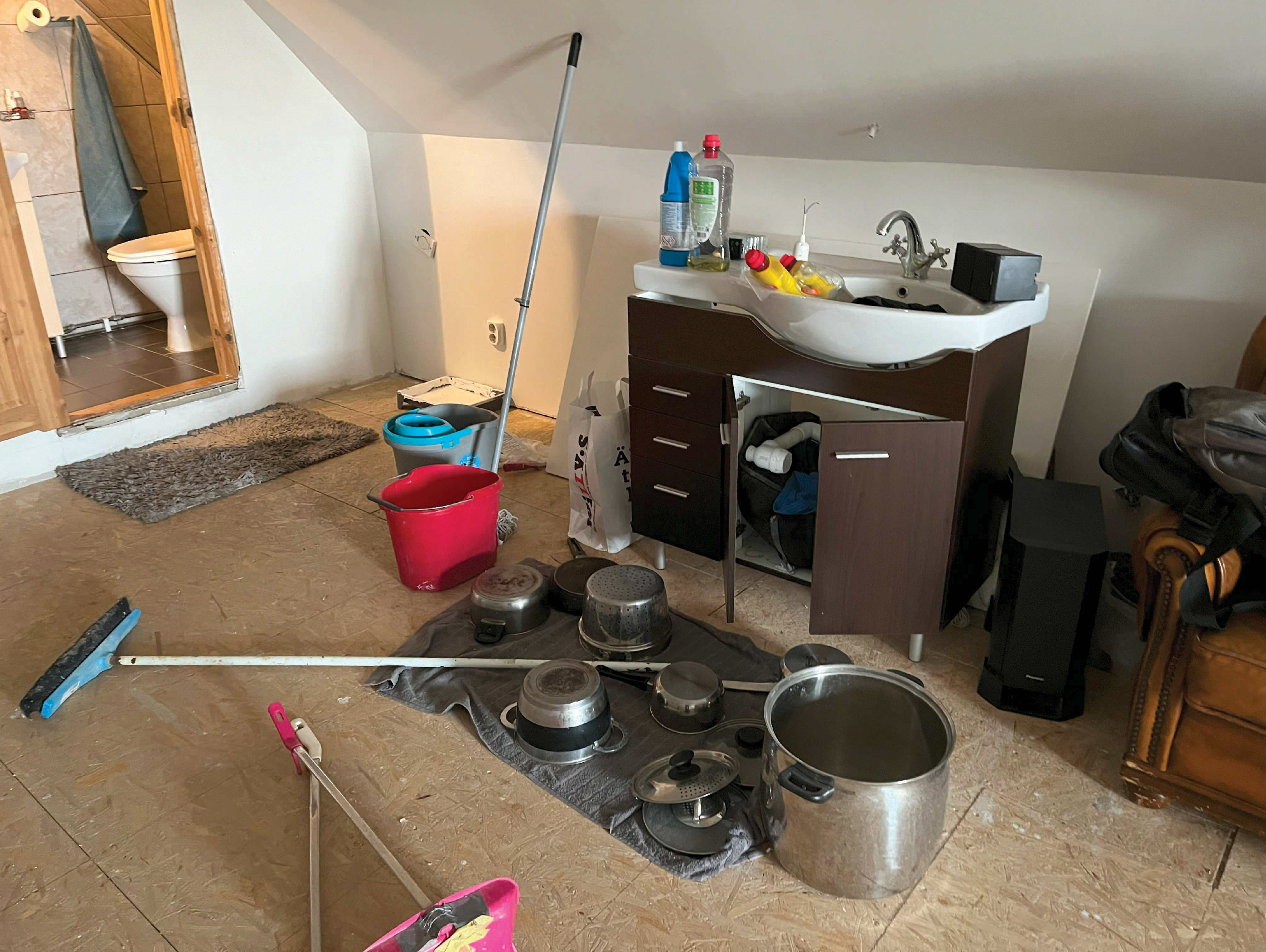 Ett tvättställ med rengöringsmedel och hinkar, kastruller torkar på en handduk på golvet, moppar och en kvast står mot väggen.