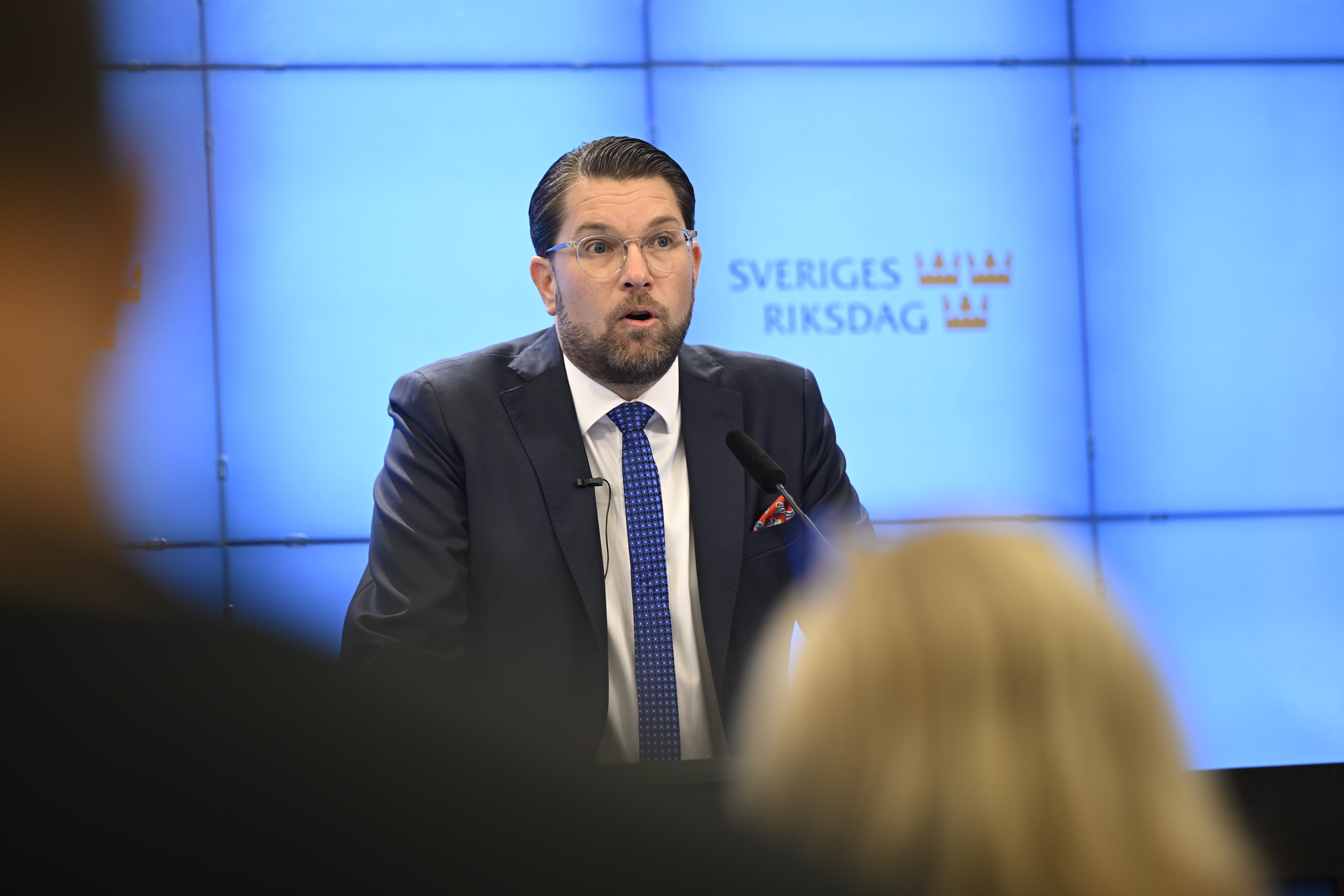 En man i kostym talar vid ett podium framför en blå skärm som visar logotypen för "Sveriges Riksdag".