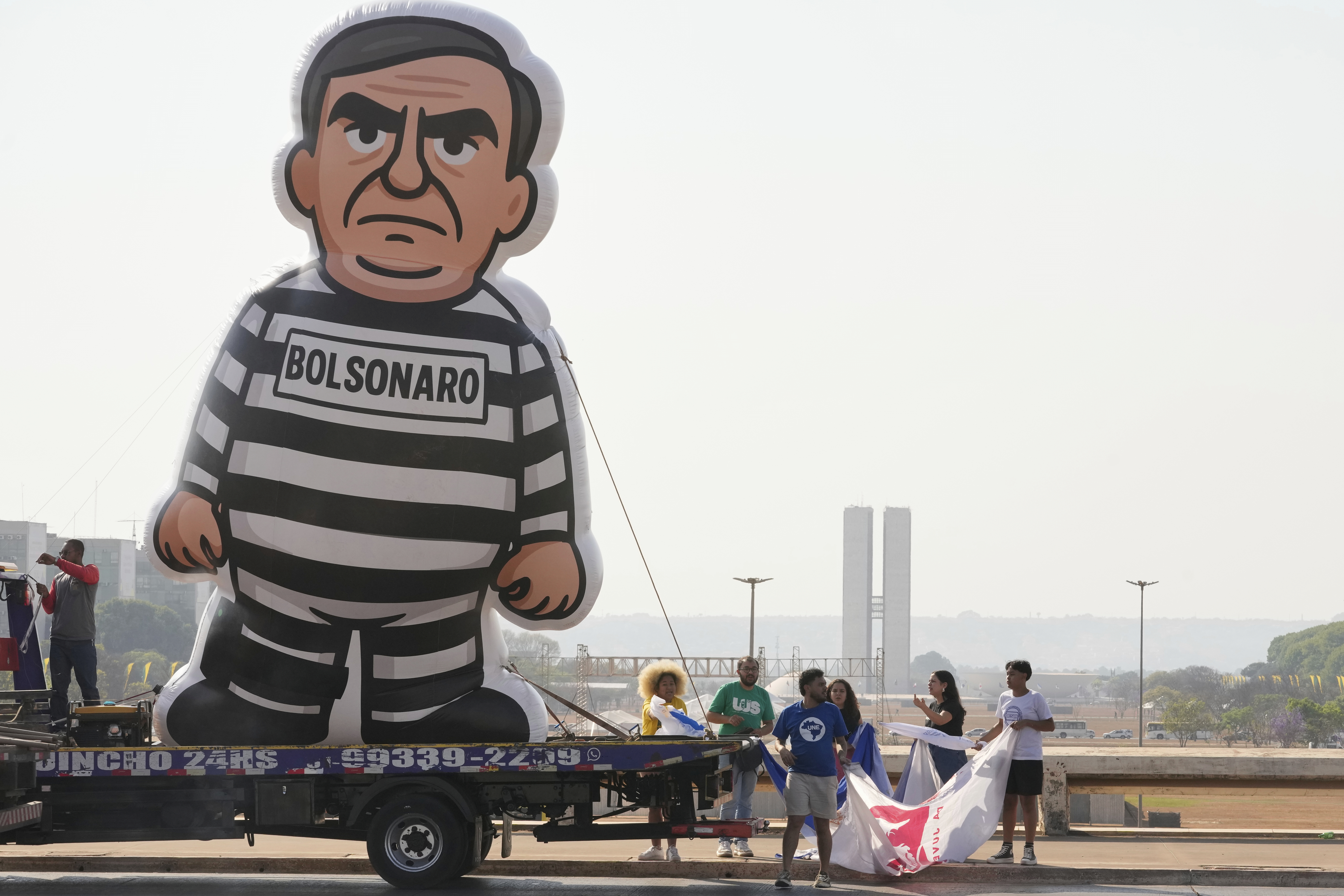En uppblåsbar figur av Bolsonaro i randig fängelsedräkt står på en lastbil, med flera personer och banderoller i närheten utomhus.