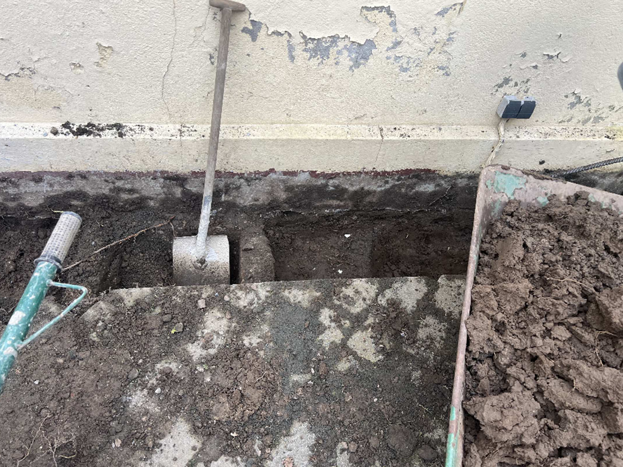 Ett delvis grävt dike löper längs en vägg. En spade och en skottkärra med jord står i närheten.