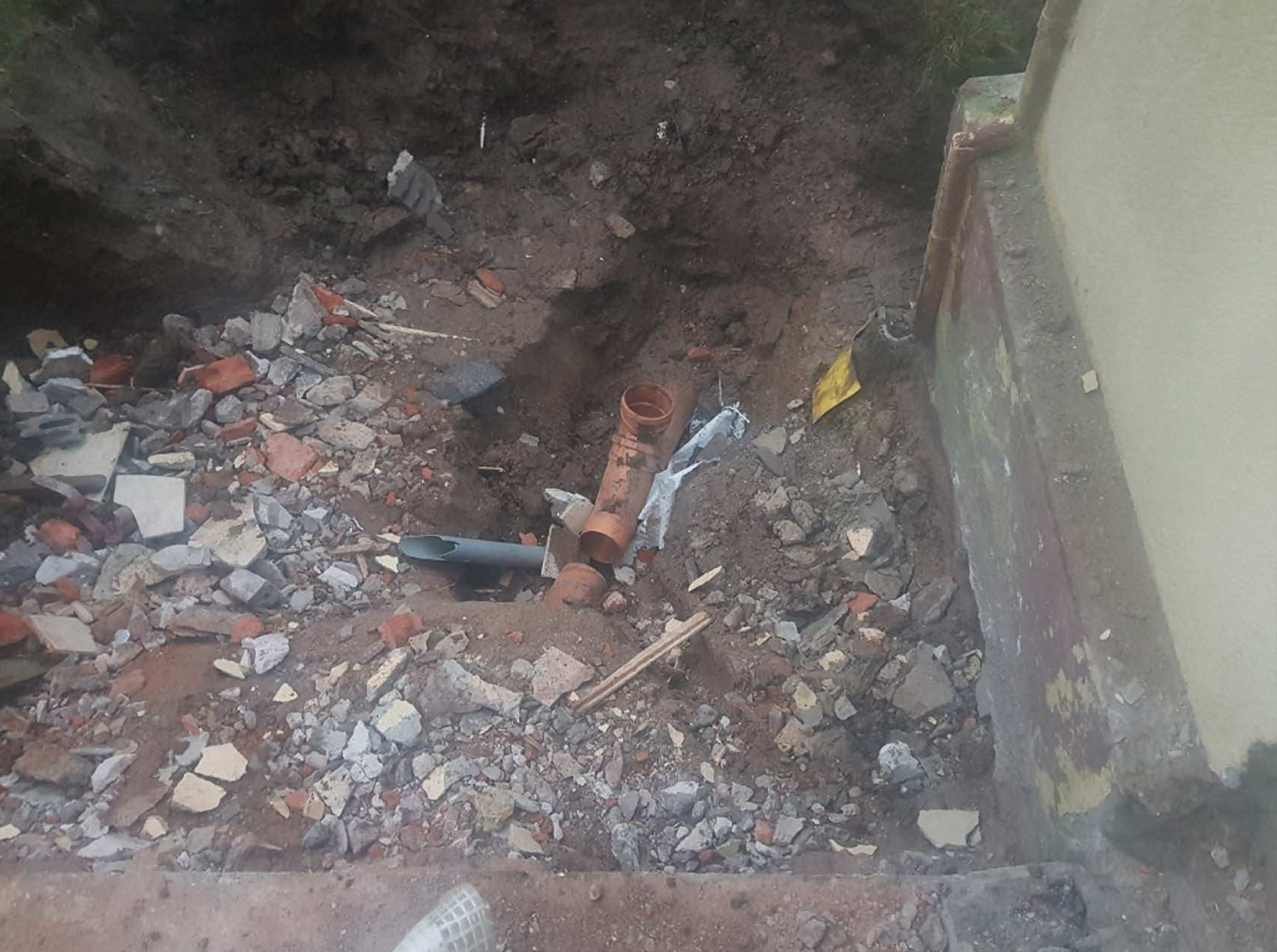 Ras och spillror omger ett skadat rör i en stor grop intill en byggnadsvägg.
