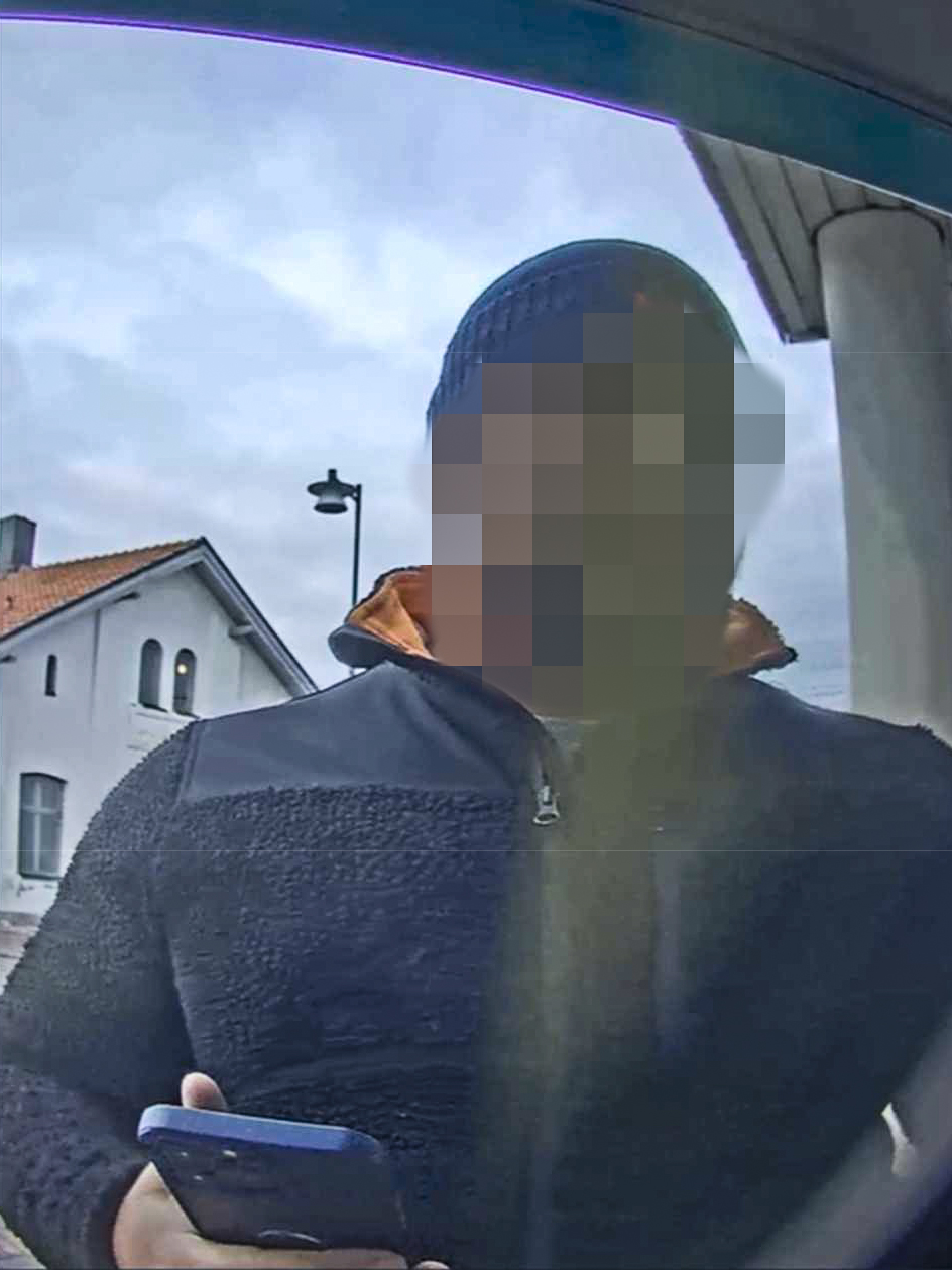 En person med pixlat ansikte står utomhus, håller en smartphone och har på sig jacka och mössa. Hus och lyktstolpe syns bakom.