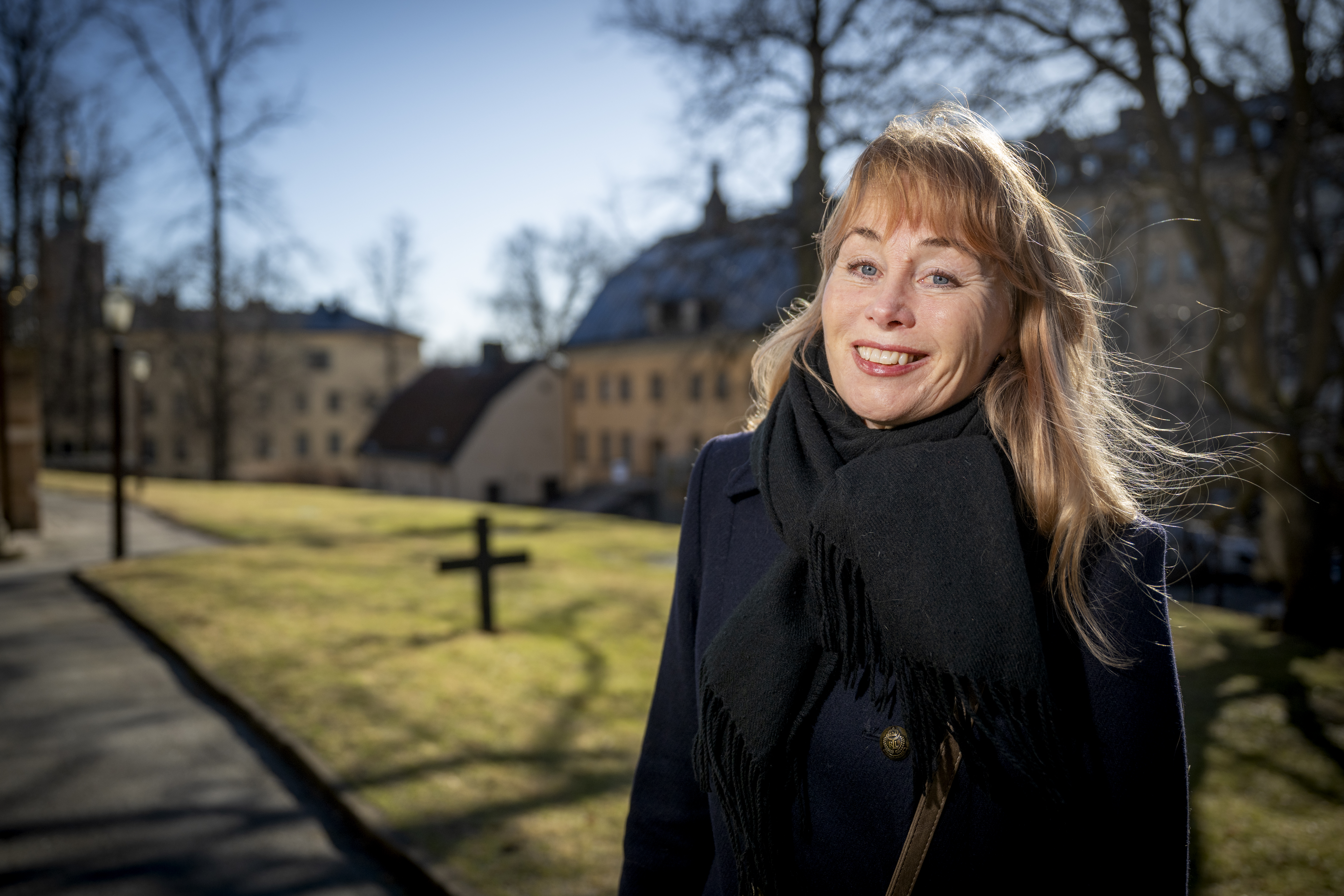En kvinna med blont hår och svart scarf står utomhus i en park med byggnader och träd utan löv i bakgrunden.