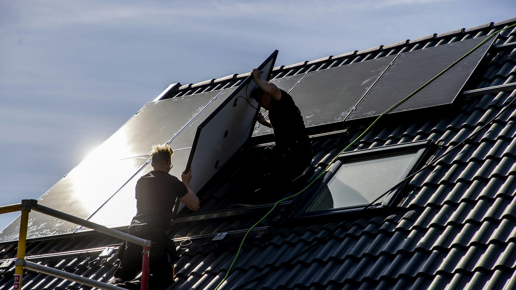 Två personer monterar solpaneler på ett tak med hjälp av byggställning och säkerhetsutrustning.