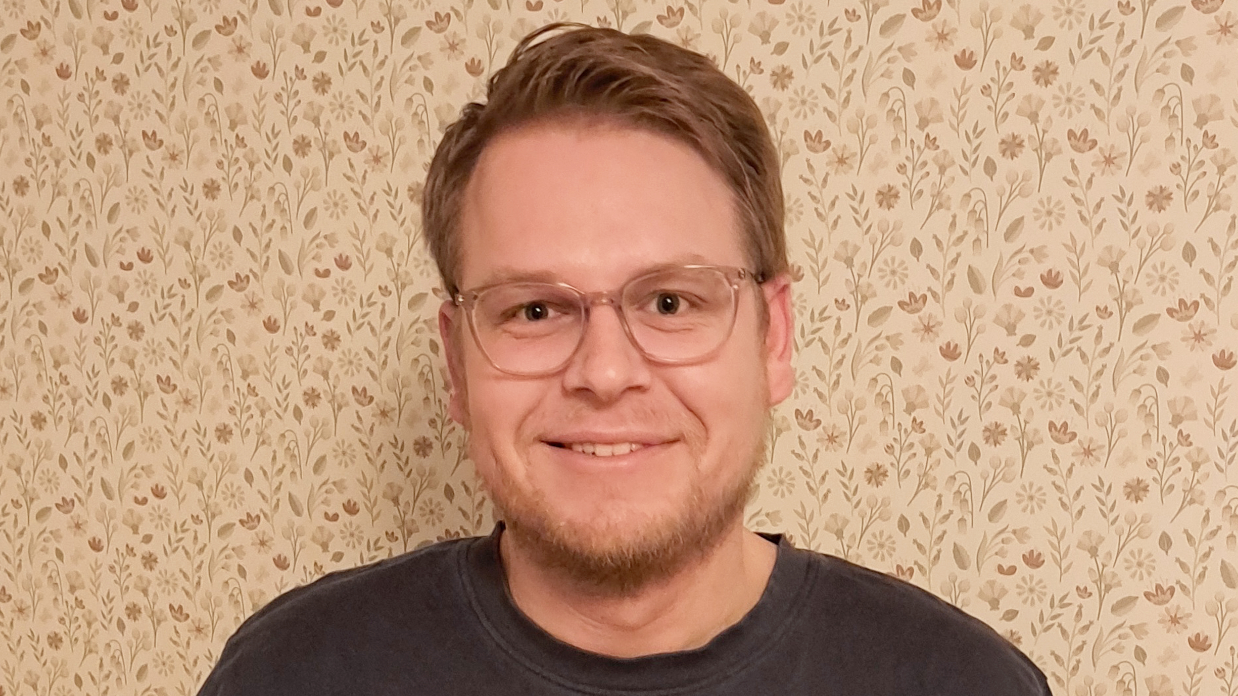 En person med kort ljusbrunt hår, glasögon och kort skägg ler och står framför en beige tapet med blommönster.