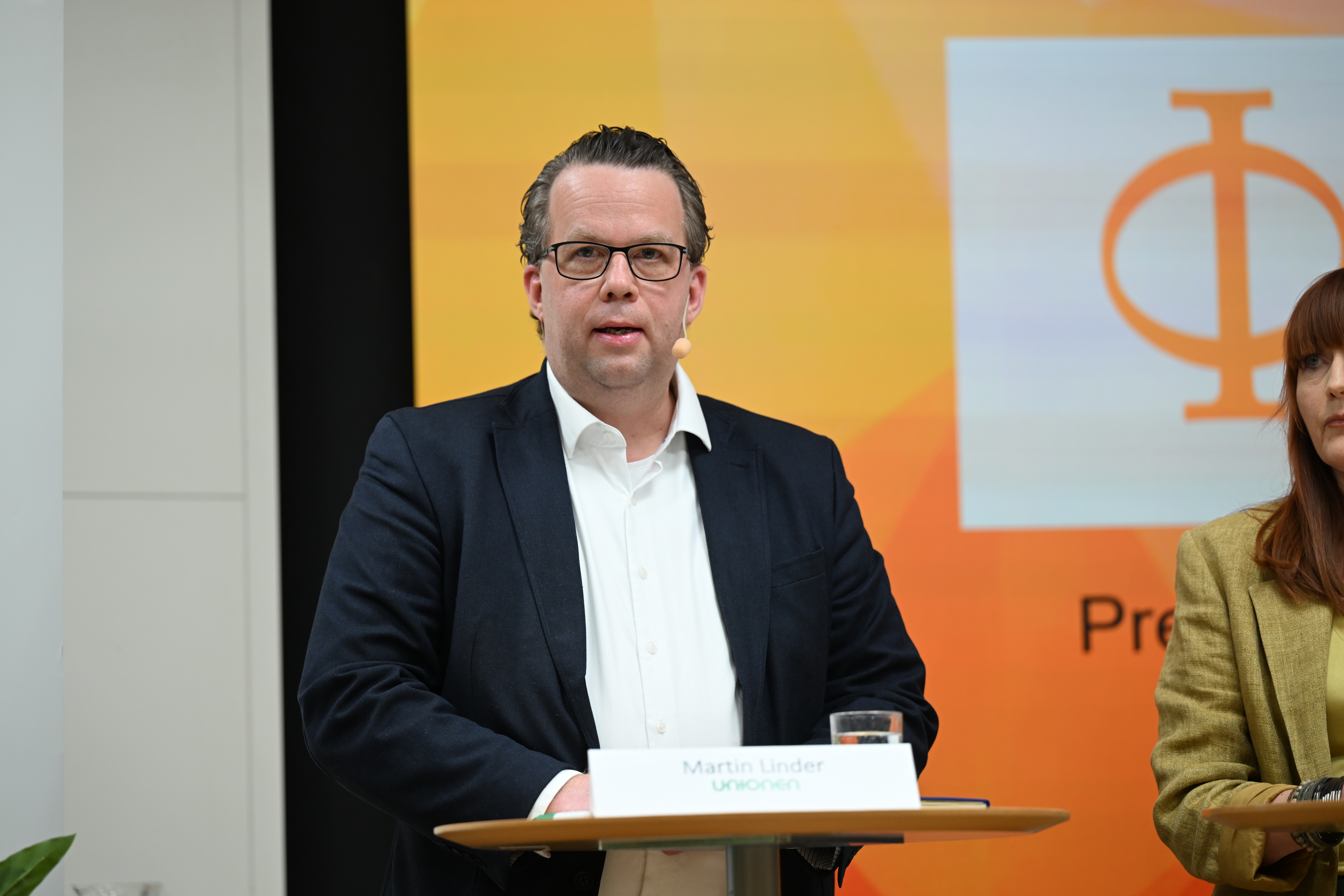 En man i kostym och glasögon talar vid ett podium med mikrofon. En kvinna syns delvis bredvid. Bakgrunden har en symbol och text.