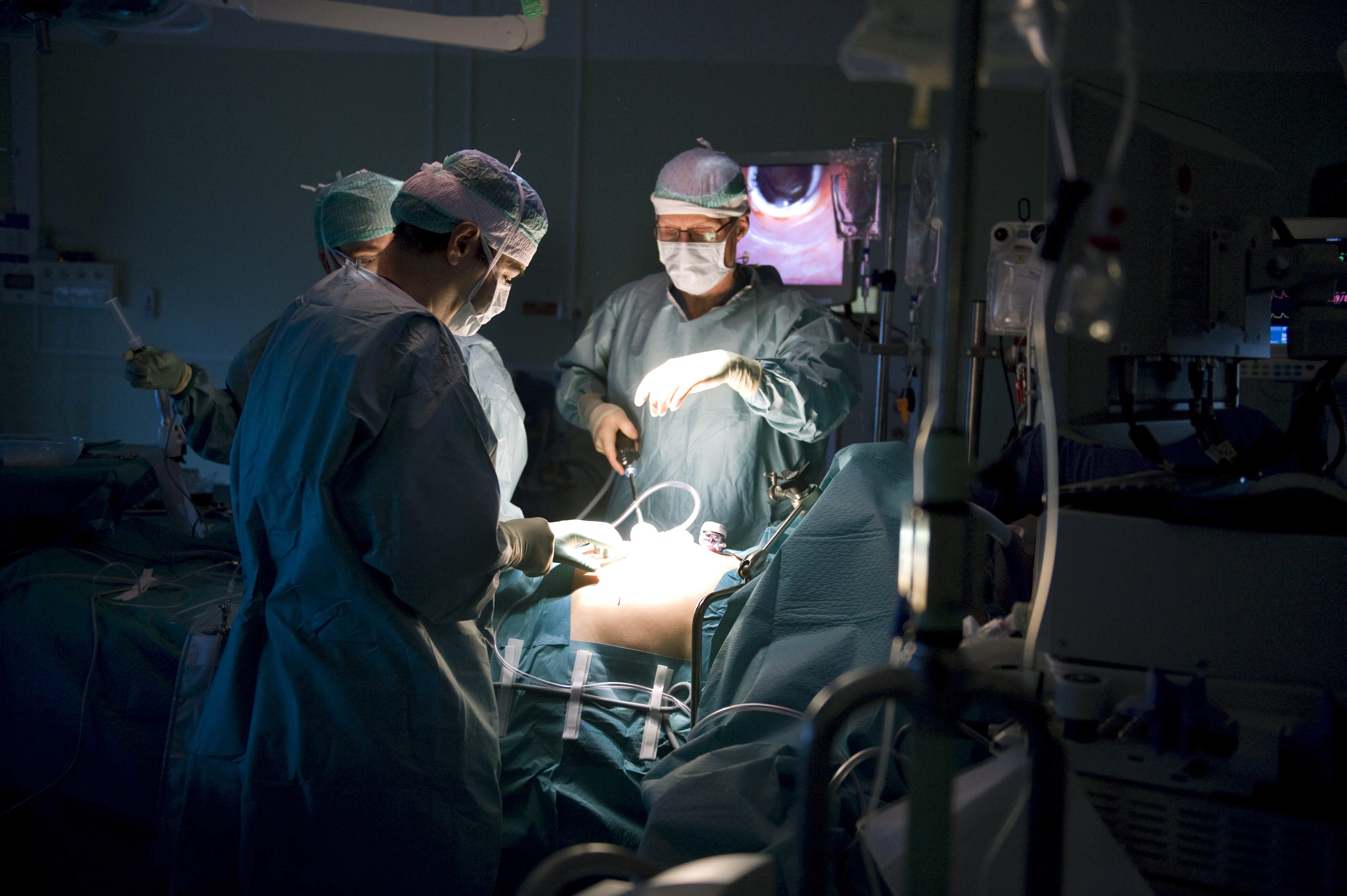 Tre kirurger i operationskläder opererar en patient i ett operationsrum och använder medicinsk utrustning och operationslampor.