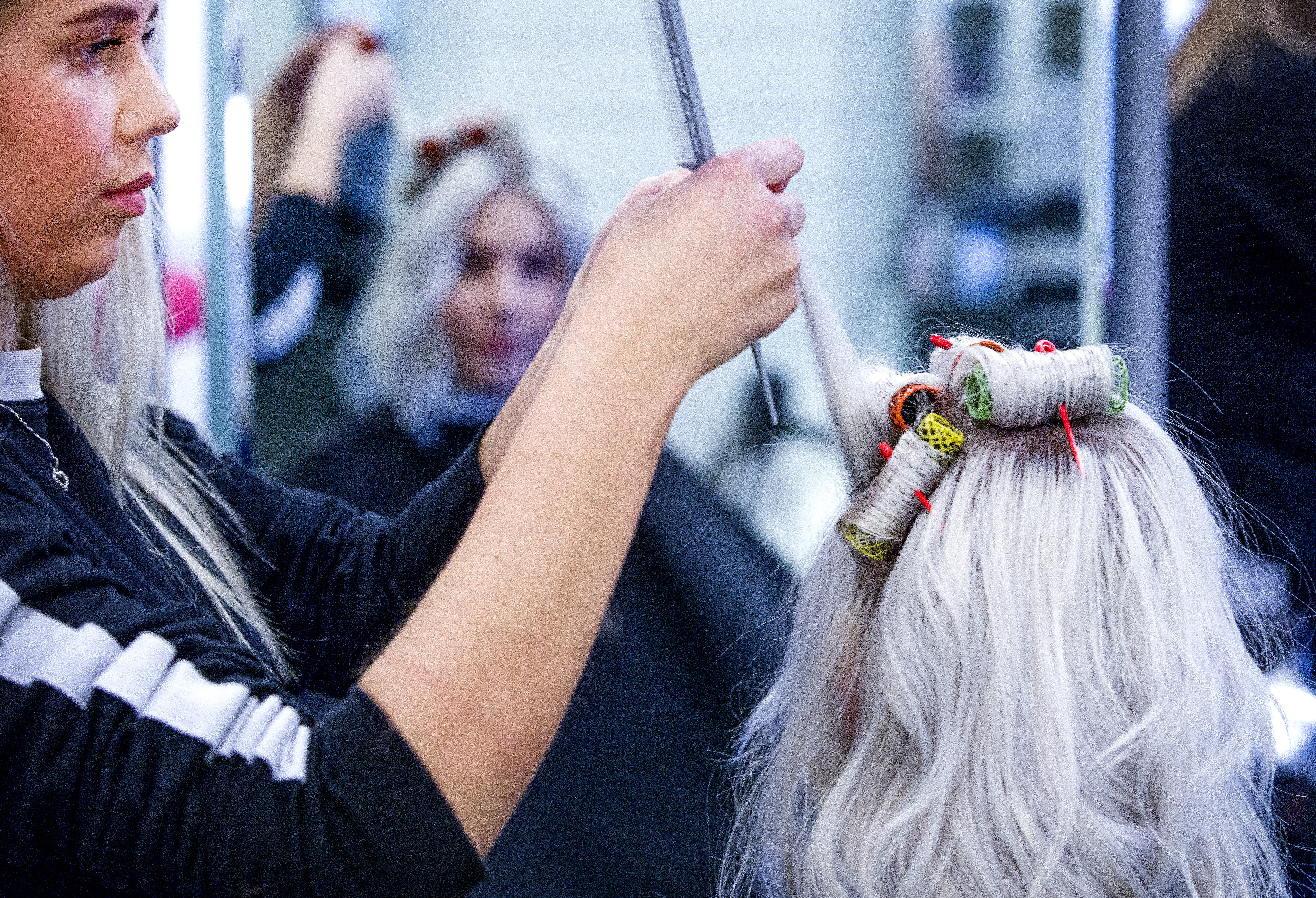 En frisör rullar upp delar av en kunds långa, blonda hår på permanentspolar, med kundens spegelbild i spegeln.