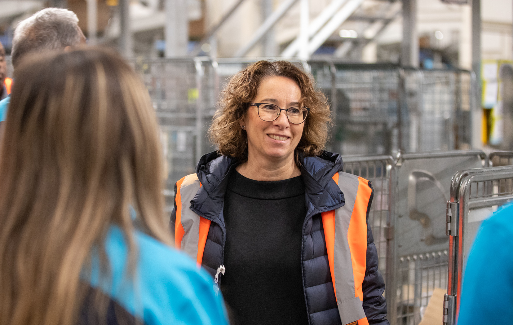 En kvinna med orange säkerhetsväst och glasögon står inomhus med metallvagnar och personer i arbetskläder omkring sig.