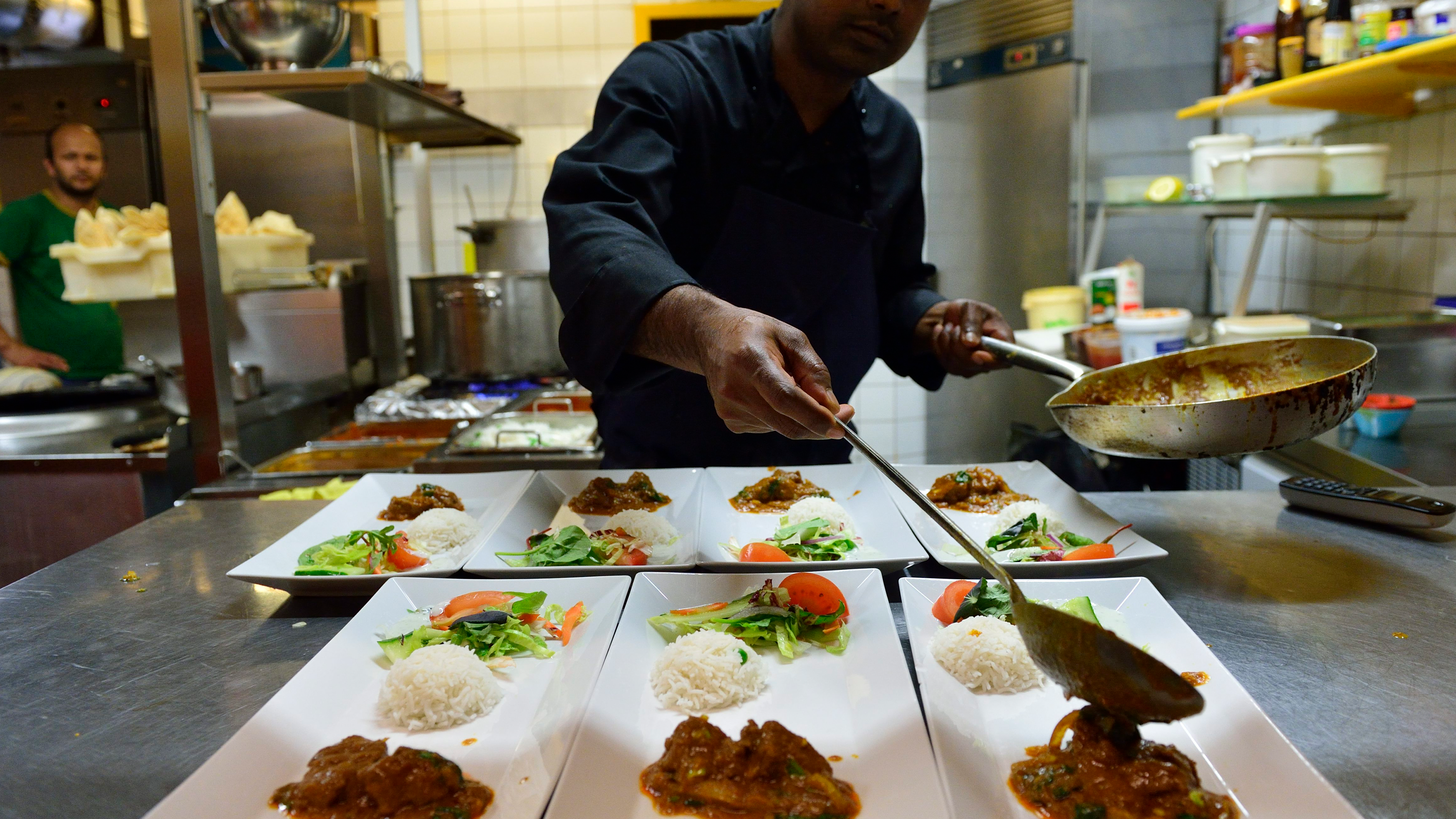 En kock lägger upp curry med ris och grönsaker på rektangulära vita tallrikar i ett restaurangkök.