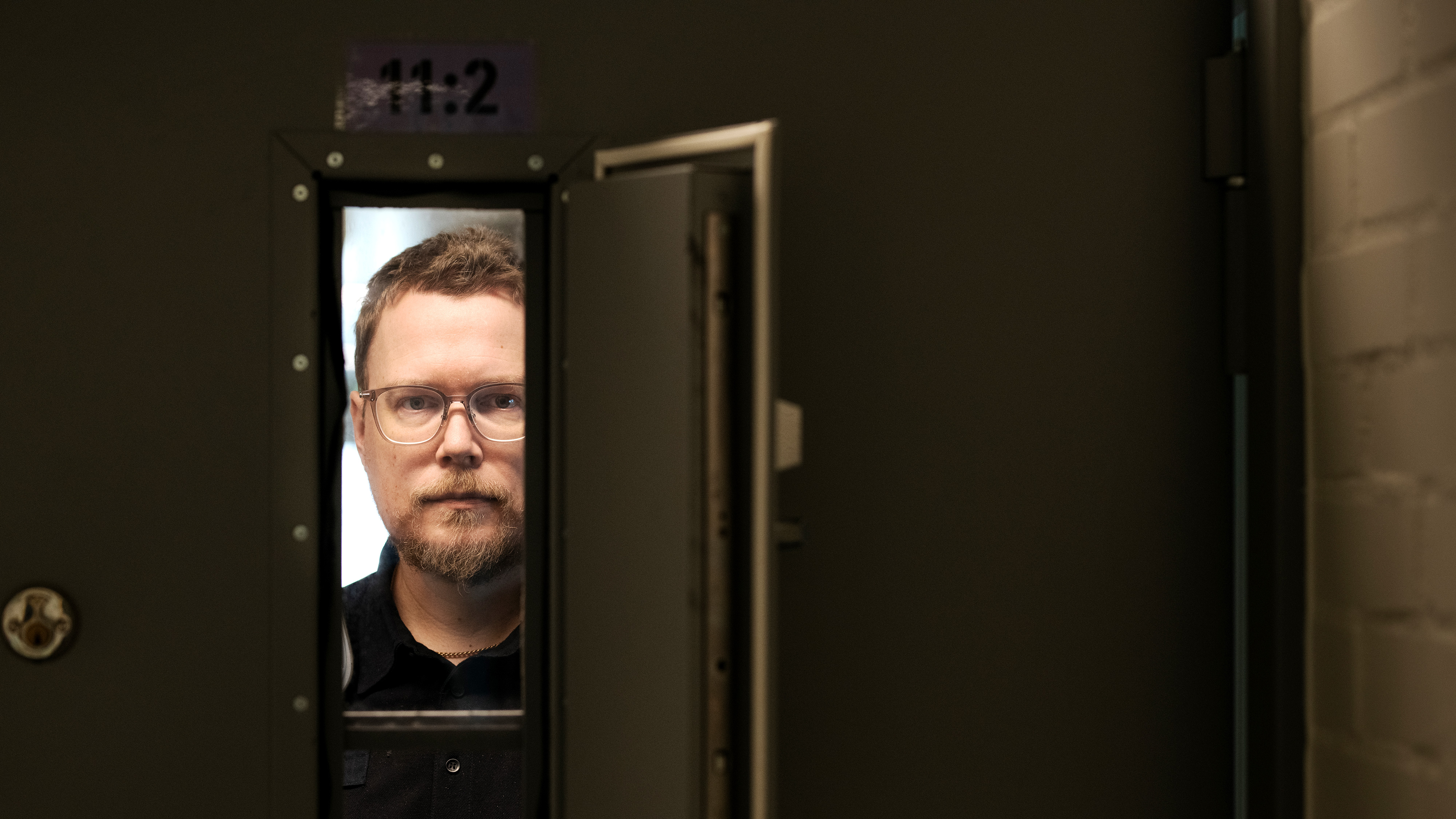 En man med glasögon tittar genom ett rektangulärt fönster i en metalldörr märkt med "11:2".