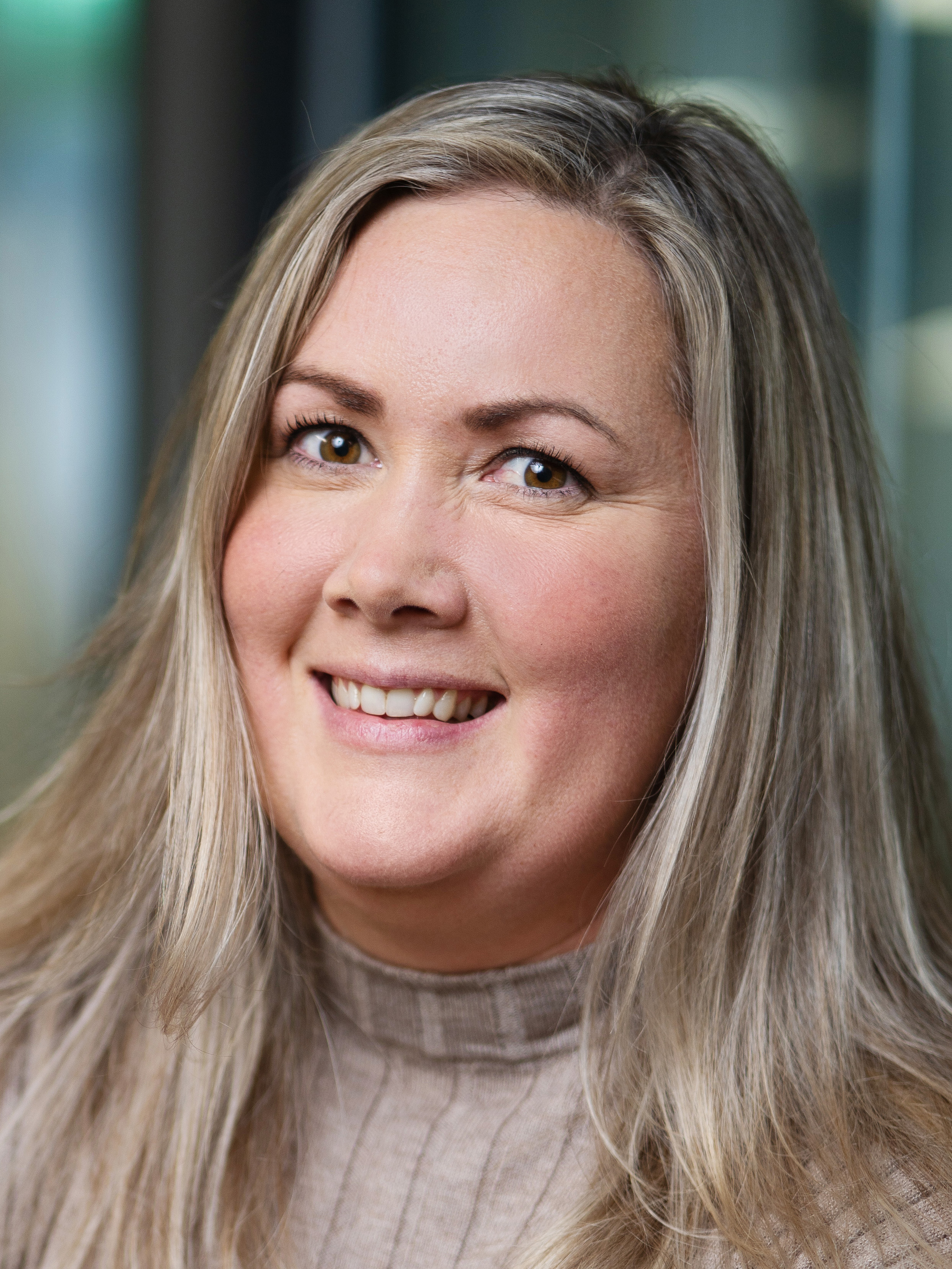 En kvinna med långt blont hår och en beige polotröja ler mot kameran i en inomhusmiljö.