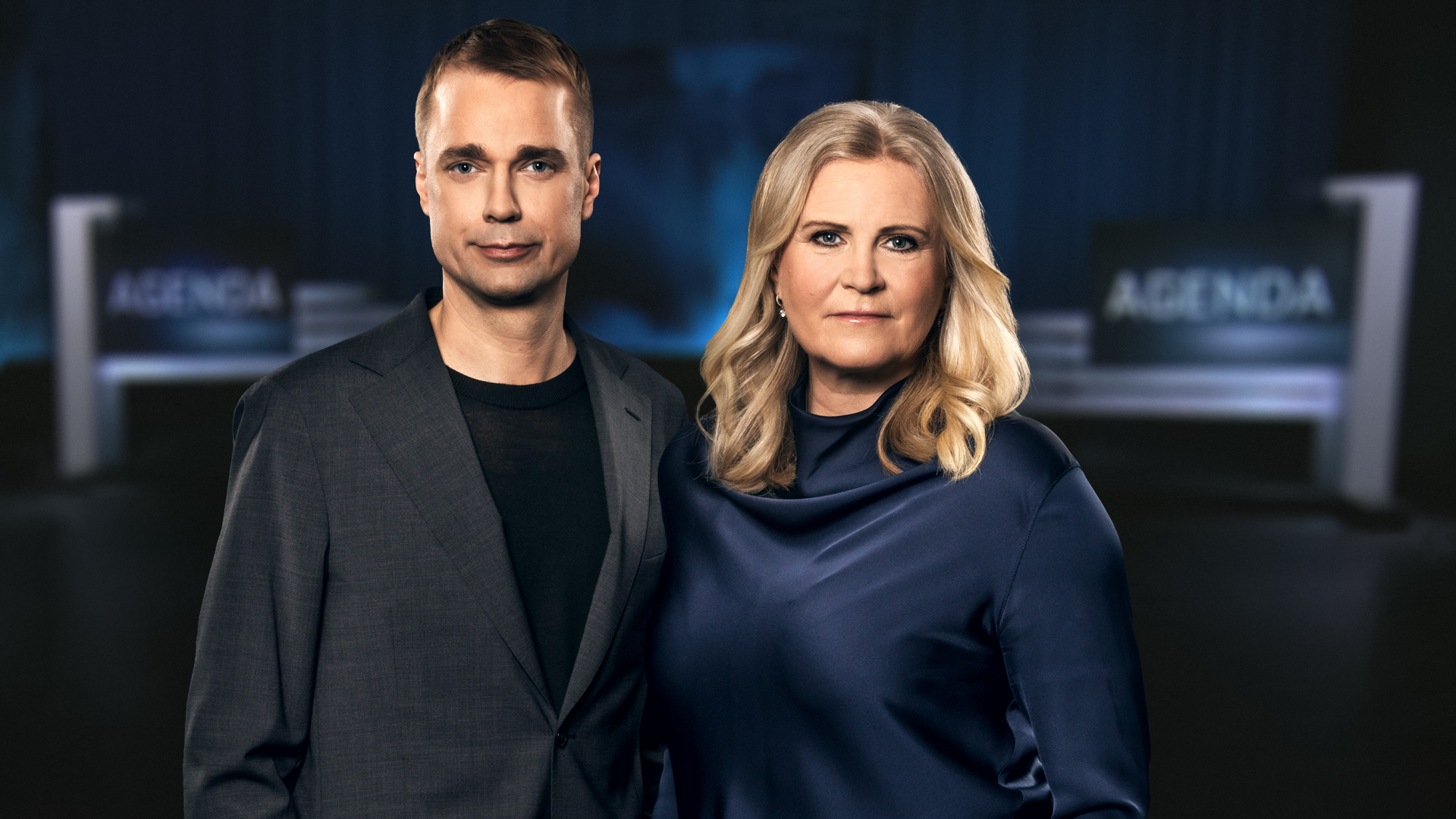 Två personer står bredvid varandra i en studio med suddiga "AGENDA"-skyltar i bakgrunden. Båda tittar framåt och har mörka kläder.