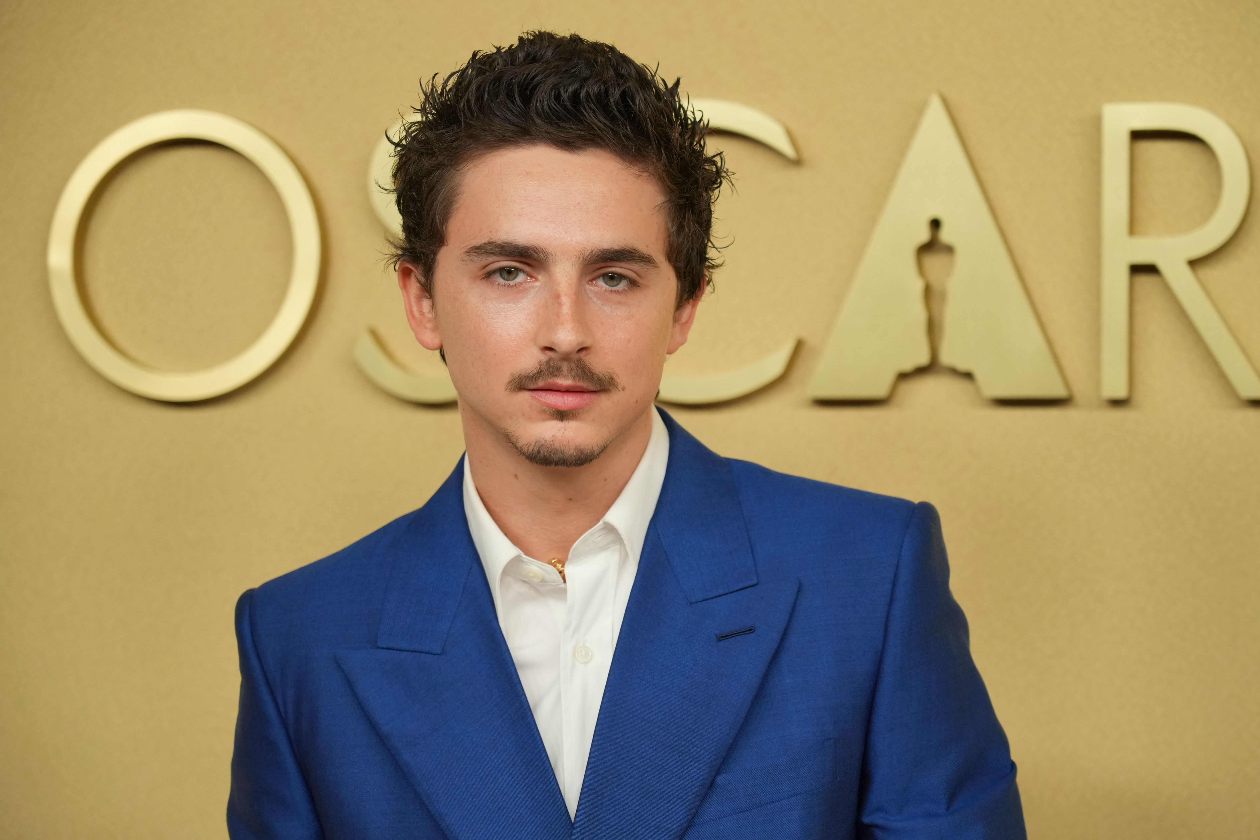 En man i blå kostym och vit skjorta står framför en guldfärgad bakgrund med ordet "OSCAR" delvis synligt.