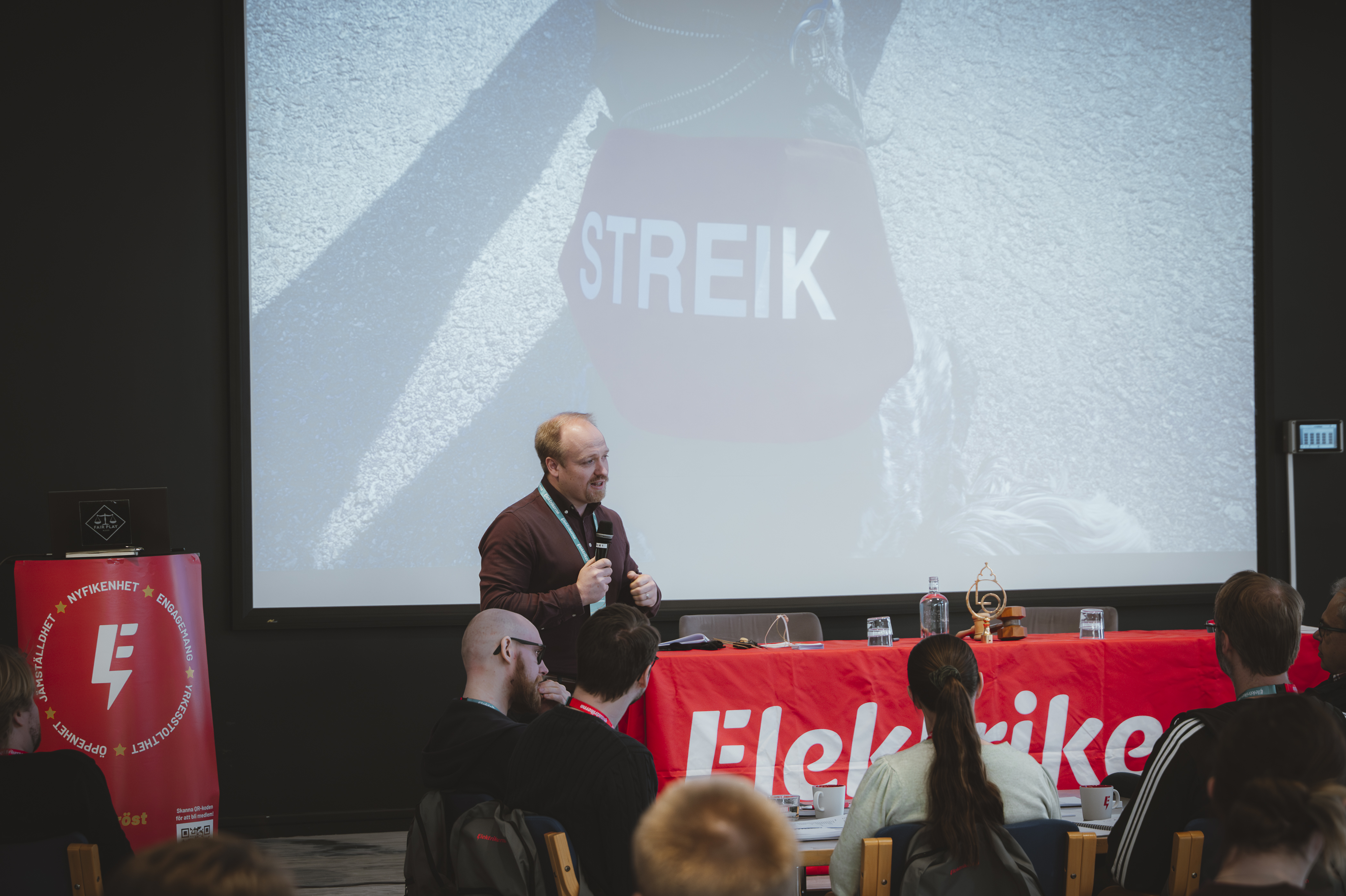 En man talar i en mikrofon vid ett podium framför en publik, med en "STREIK"-skylt och en "Elektriker"-banner bakom.