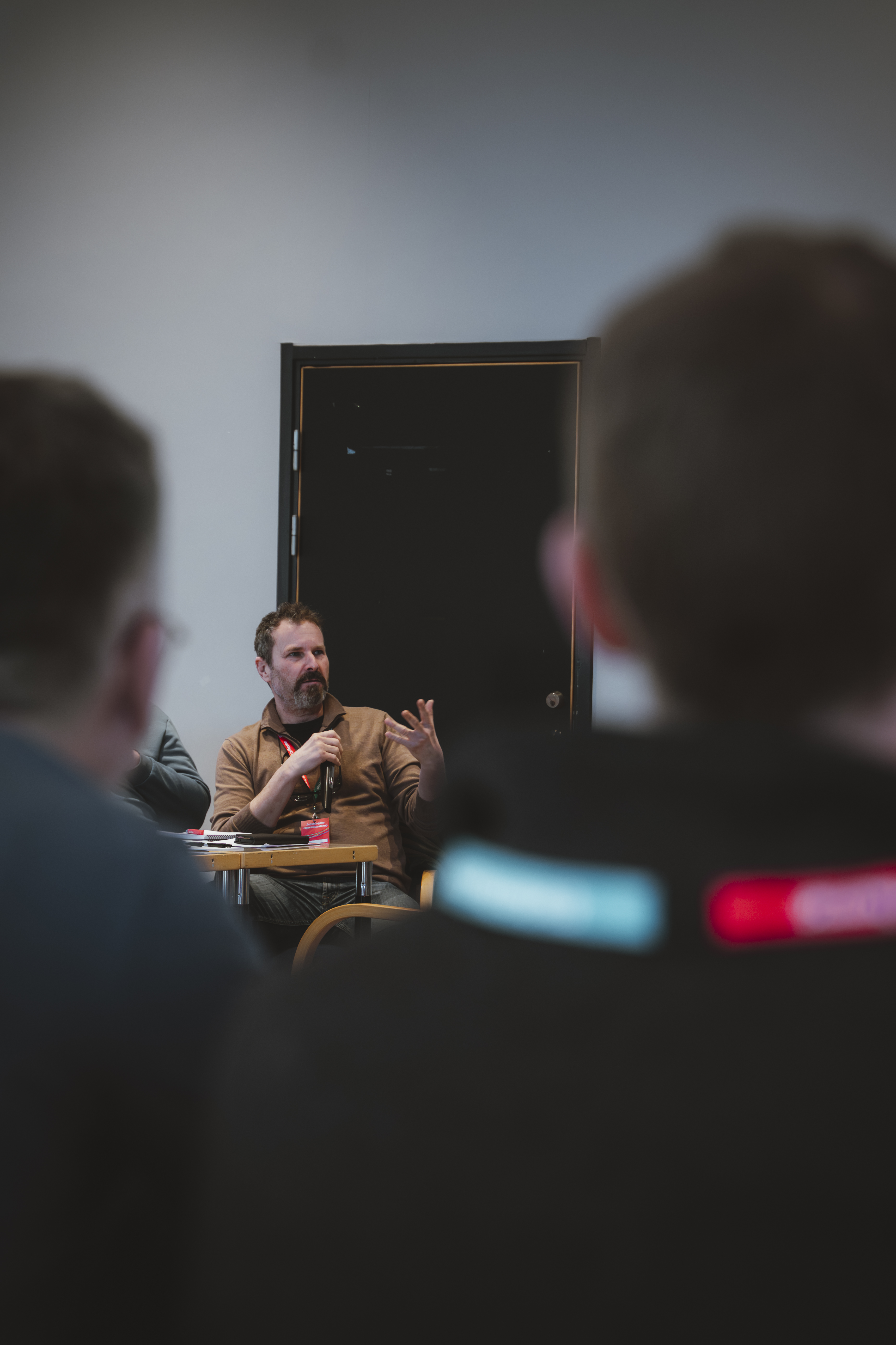 En man sitter vid ett bord och pratar samt gestikulerar med händerna, medan två personer i förgrunden lyssnar och tittar på honom.