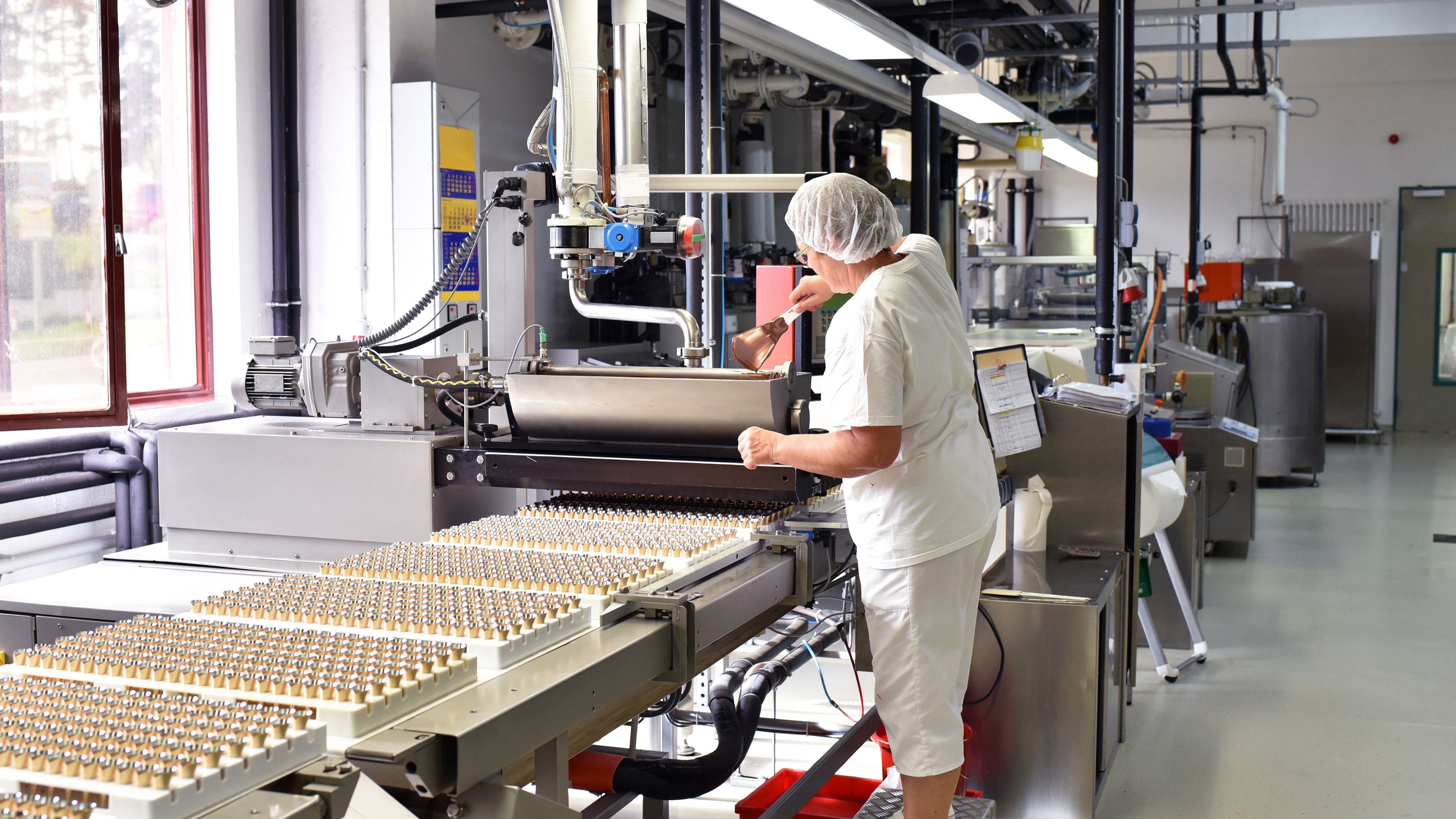 En arbetare i vit uniform och hårnät hanterar maskiner i en fabrik och har uppsikt över små flaskor på ett transportband.