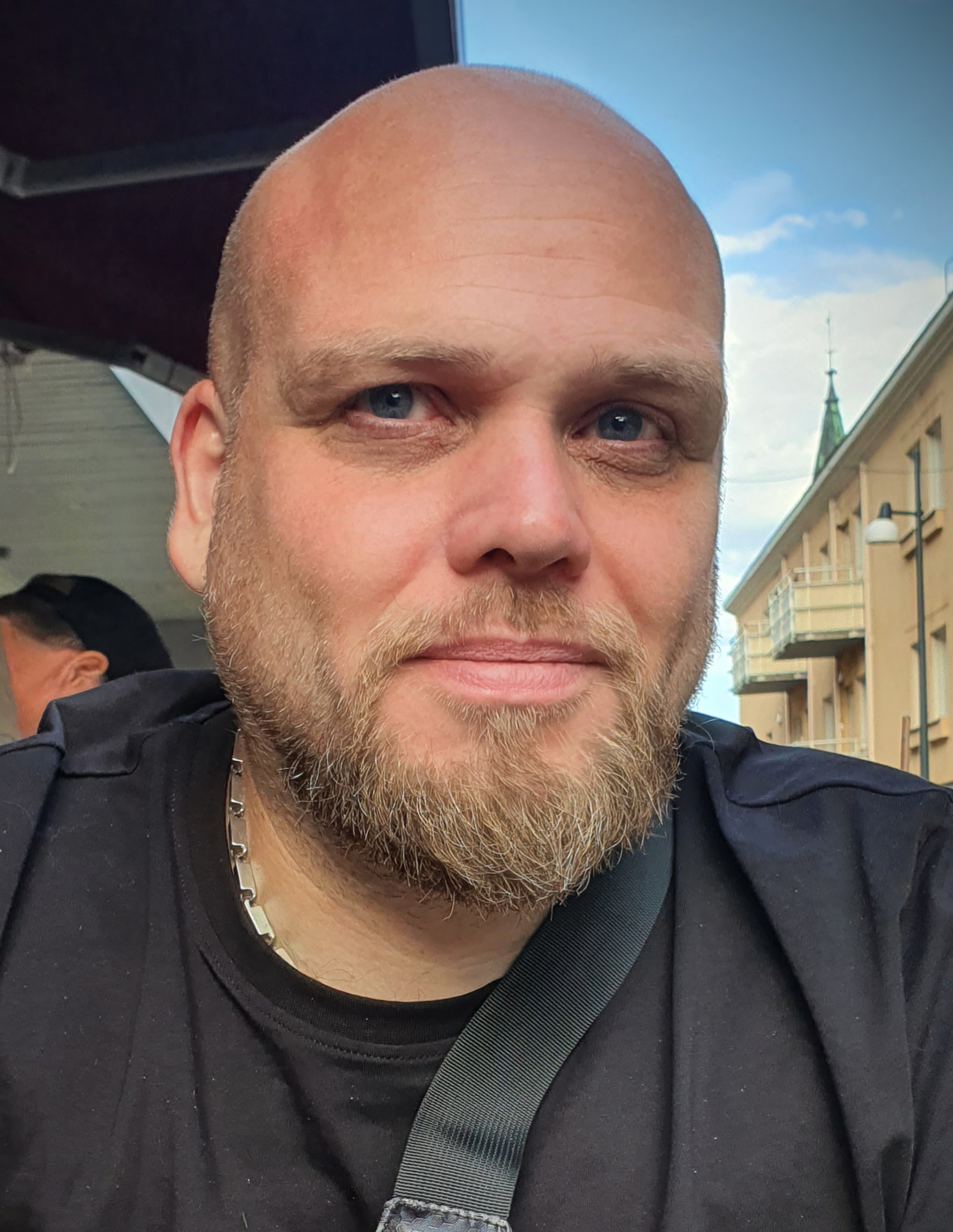 Fredrik Rosenberg i skägg och rakat huvud och kedjehalsband sitter utomhus och tittar mot kameran; byggnader och himmel i bakgrunden.