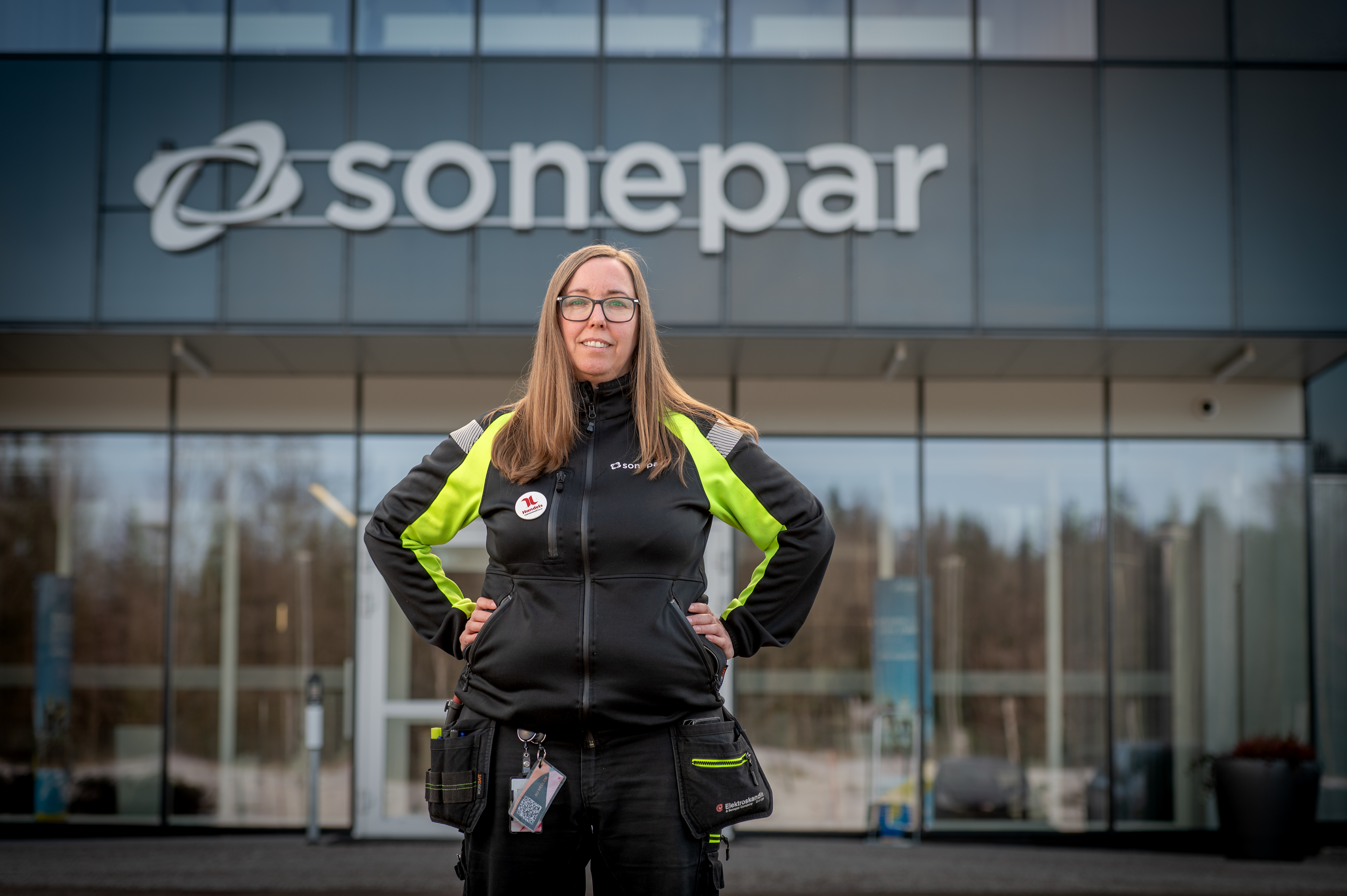 En kvinna i svart och gul arbetsjacka står med händerna på höfterna framför en byggnad med skylten "Sonepar" ovanförför ingången.