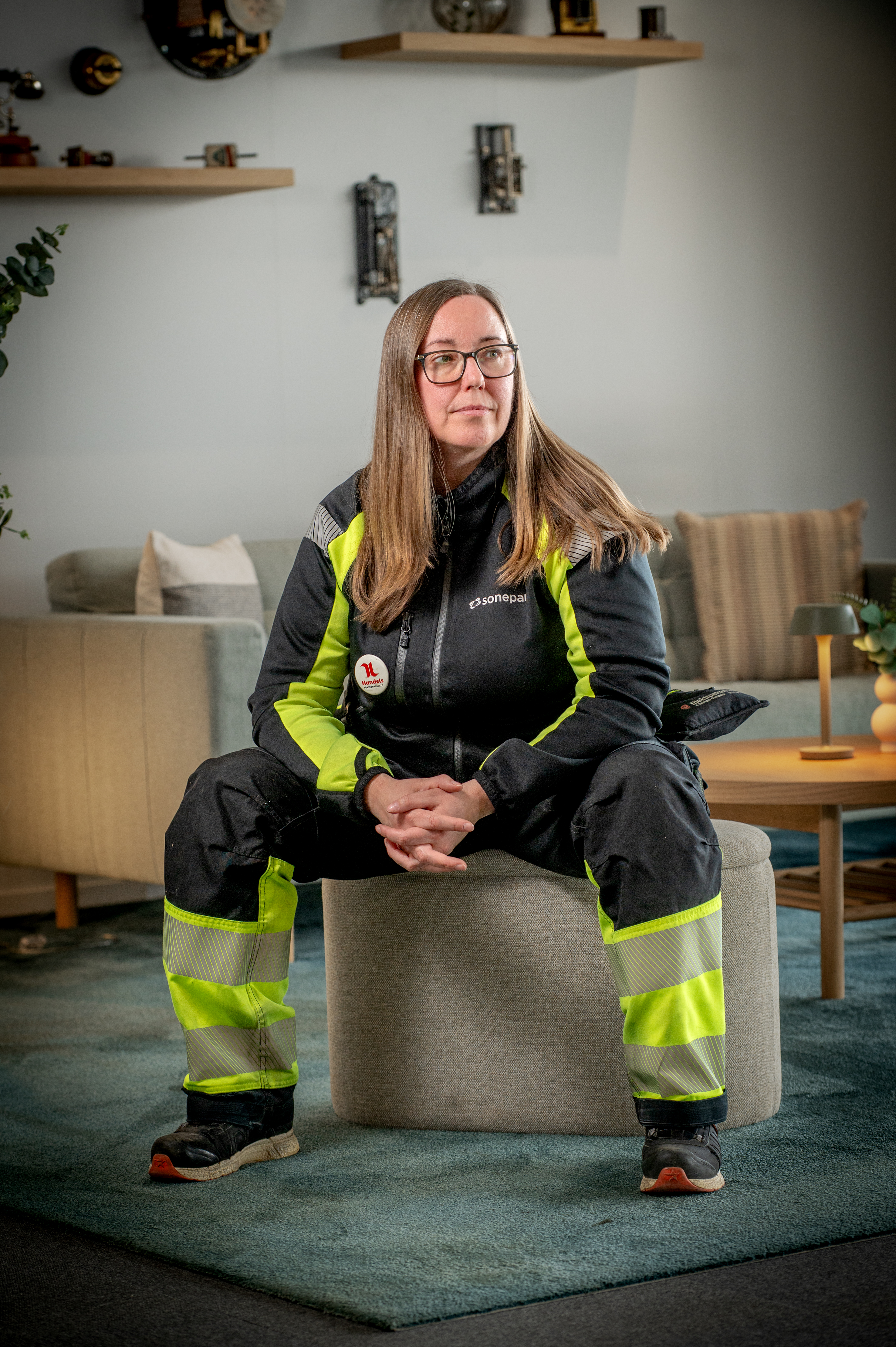 En person med långt hår och glasögon sitter på en rund sittpuff, iförd svart och gul arbetsdräkt i ett modernt vardagsrum.