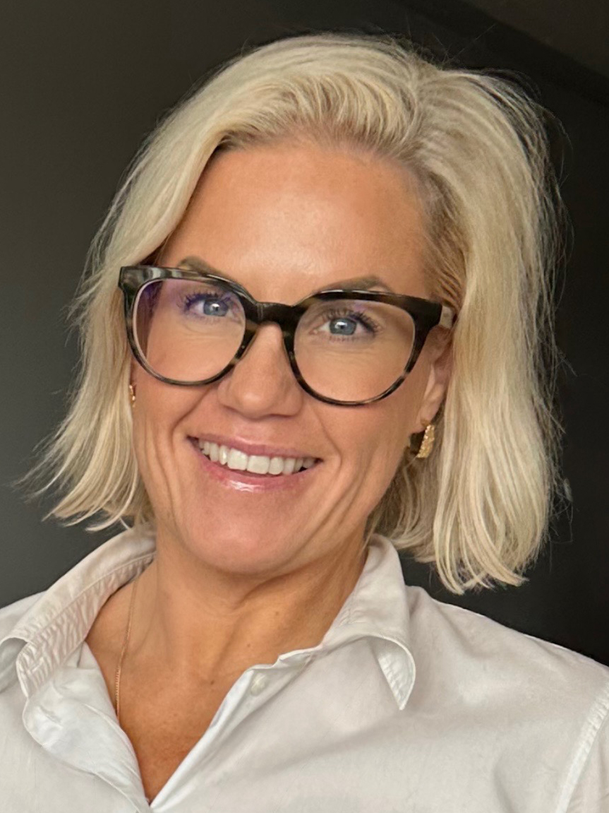 Helen Danmo med kort blont hår, svarta glasögon och vit skjorta ler mot kameran framför en mörk bakgrund.