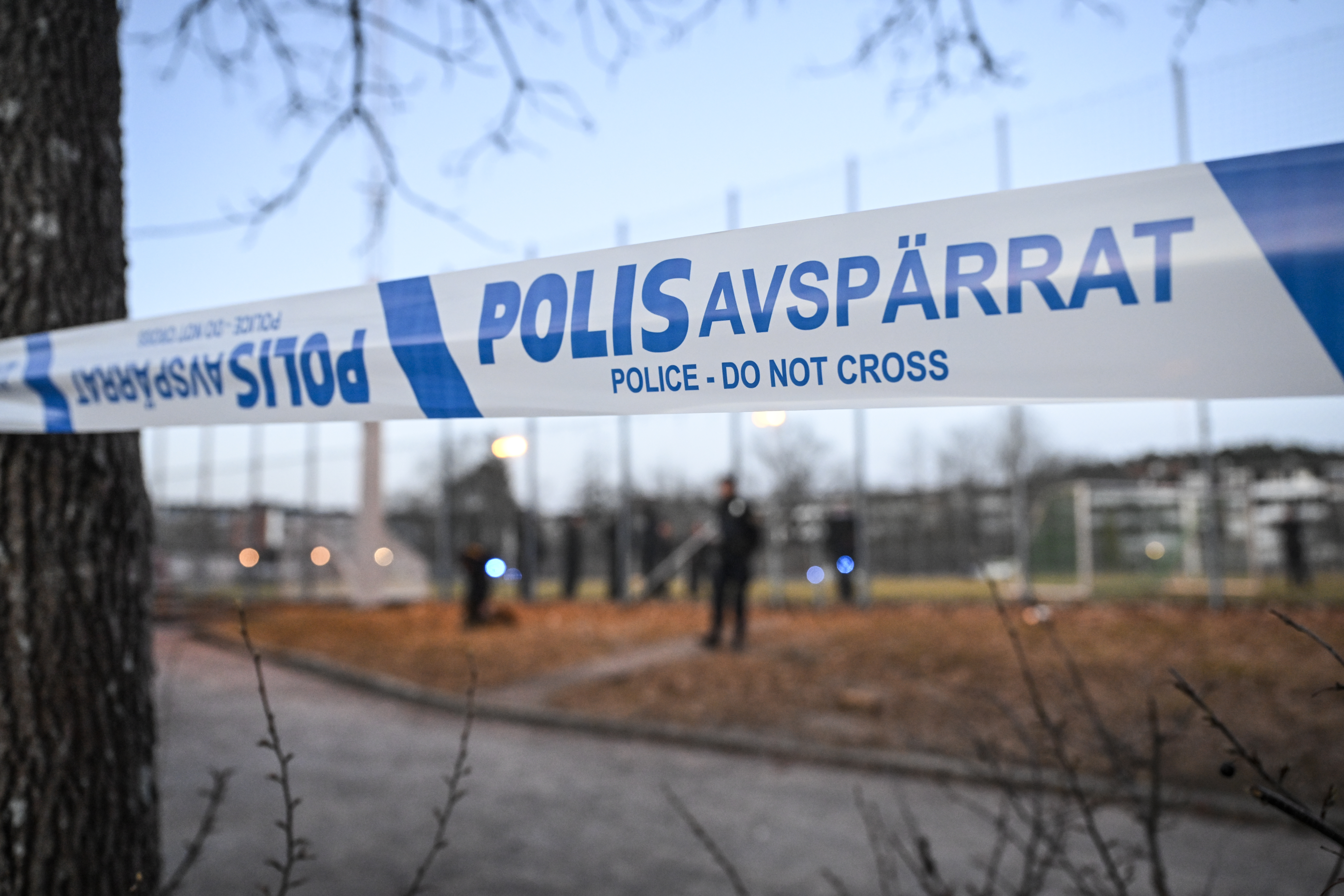 Polisavspärrning och poliser i ofokus i bakgrunden vid ett skogsbryn vid ett idrottsfält.