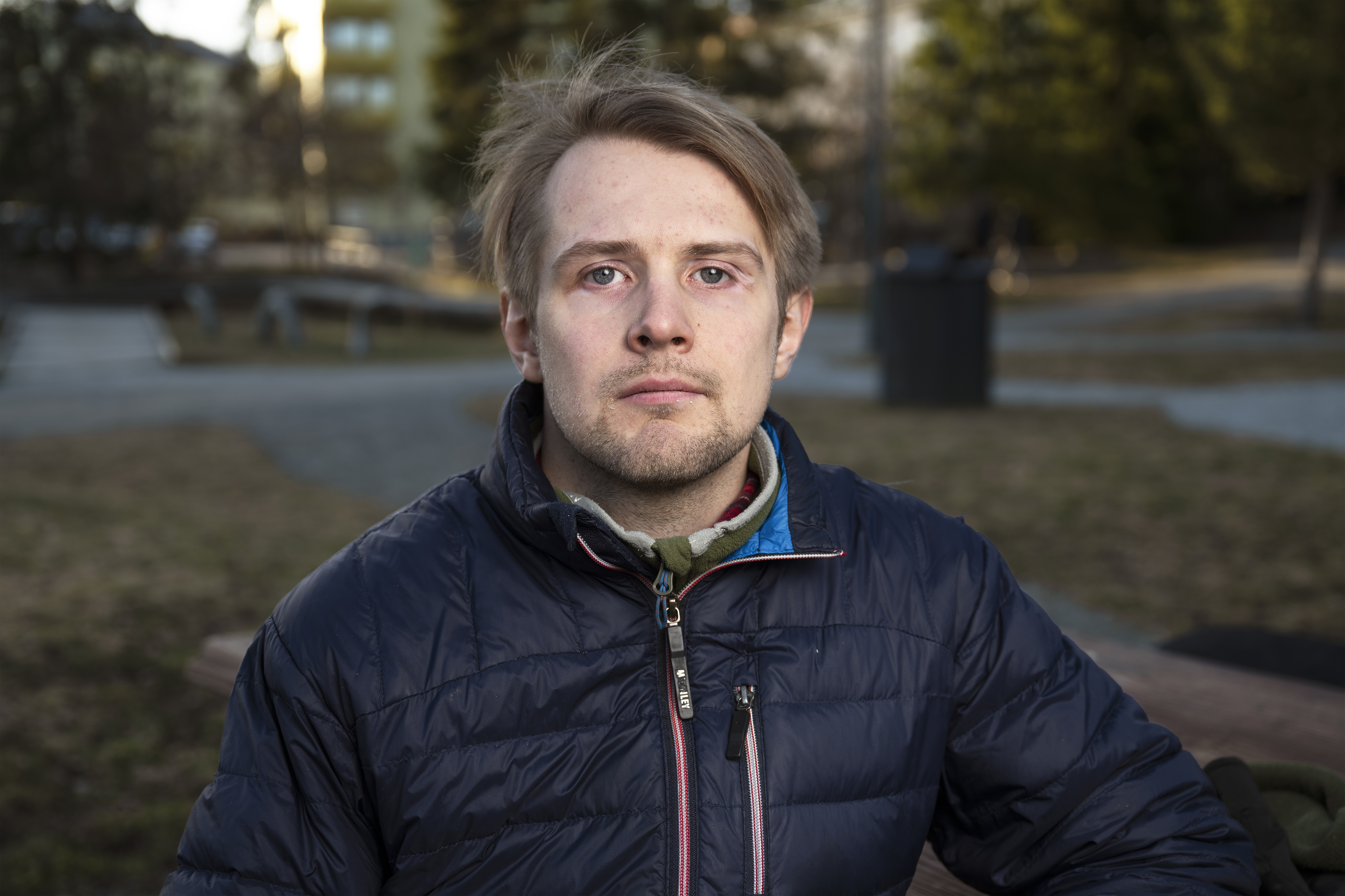Tobias, med ljusbrunt hår och mörkblå jacka, sitter på en bänk utomhus och stirrar in i kameran. Du kan se träd och byggnader i bakgrunden.
