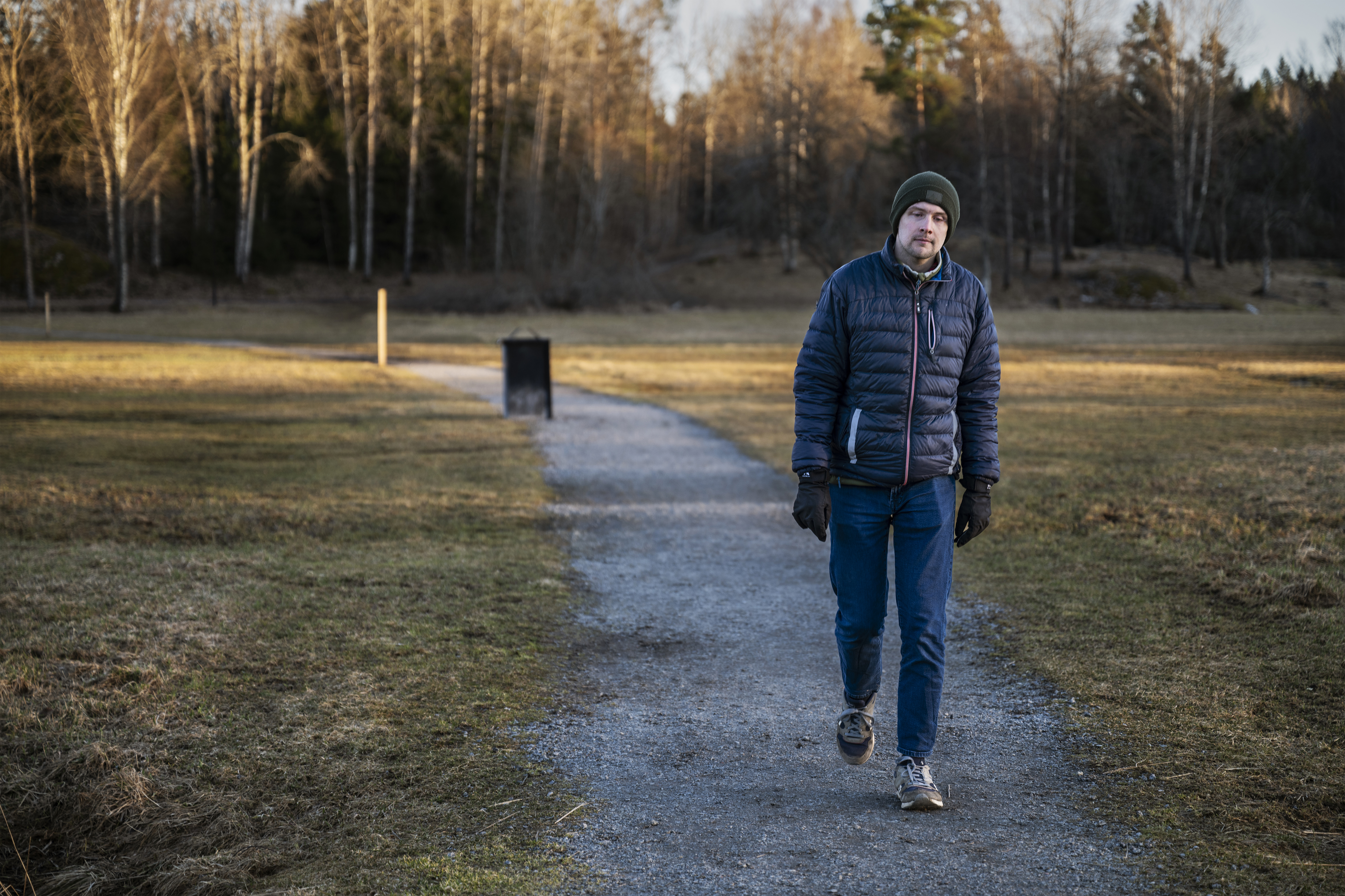 Tobias med blå jacka, mössa och handskar går ensam på en grusväg i en park med träd utan löv och torrt gräs.