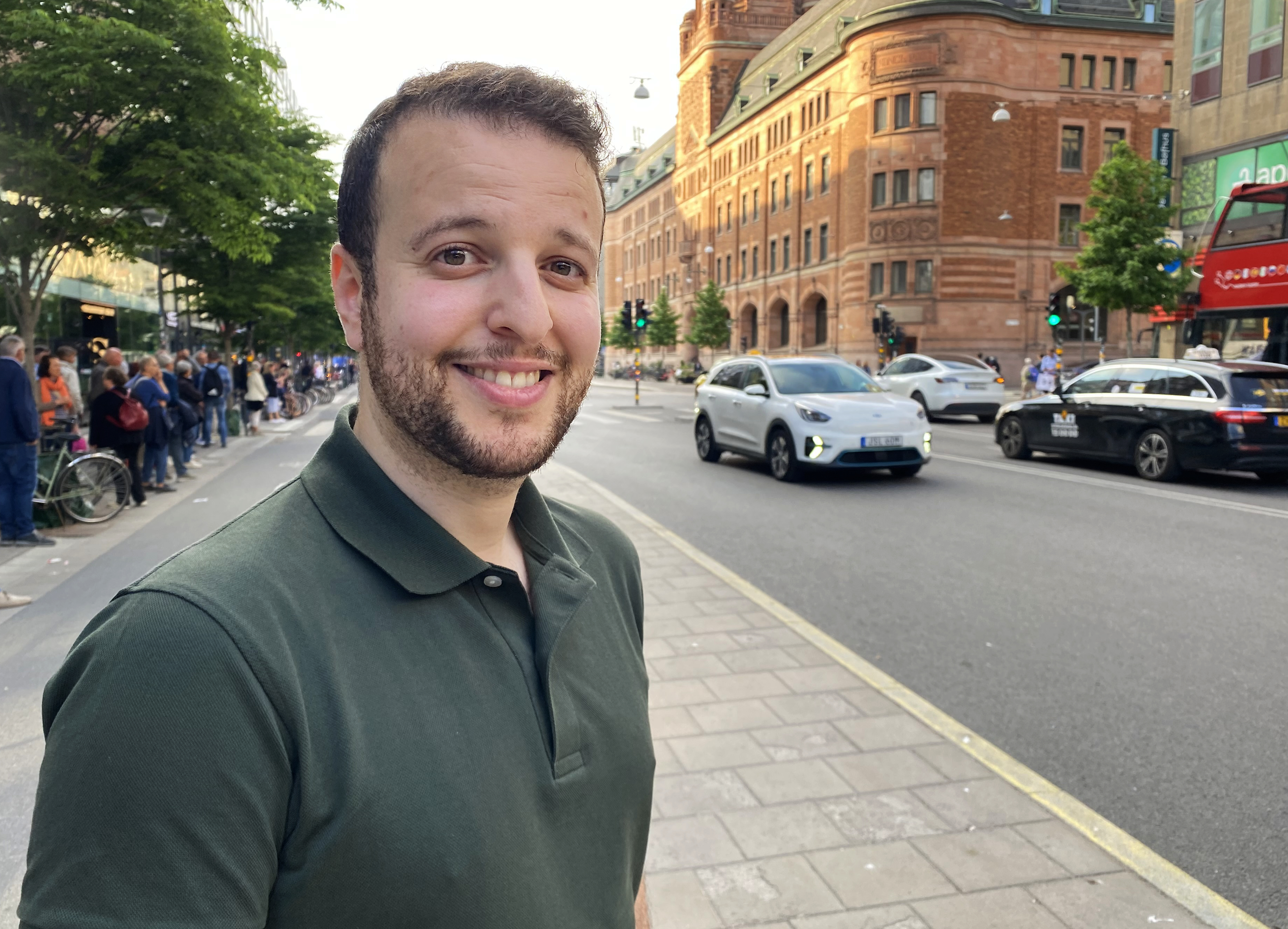 En man med mörkgrön pikétröja står på en trottoar i en stad och ler mot kameran; bilar och människor syns bredvid honom.