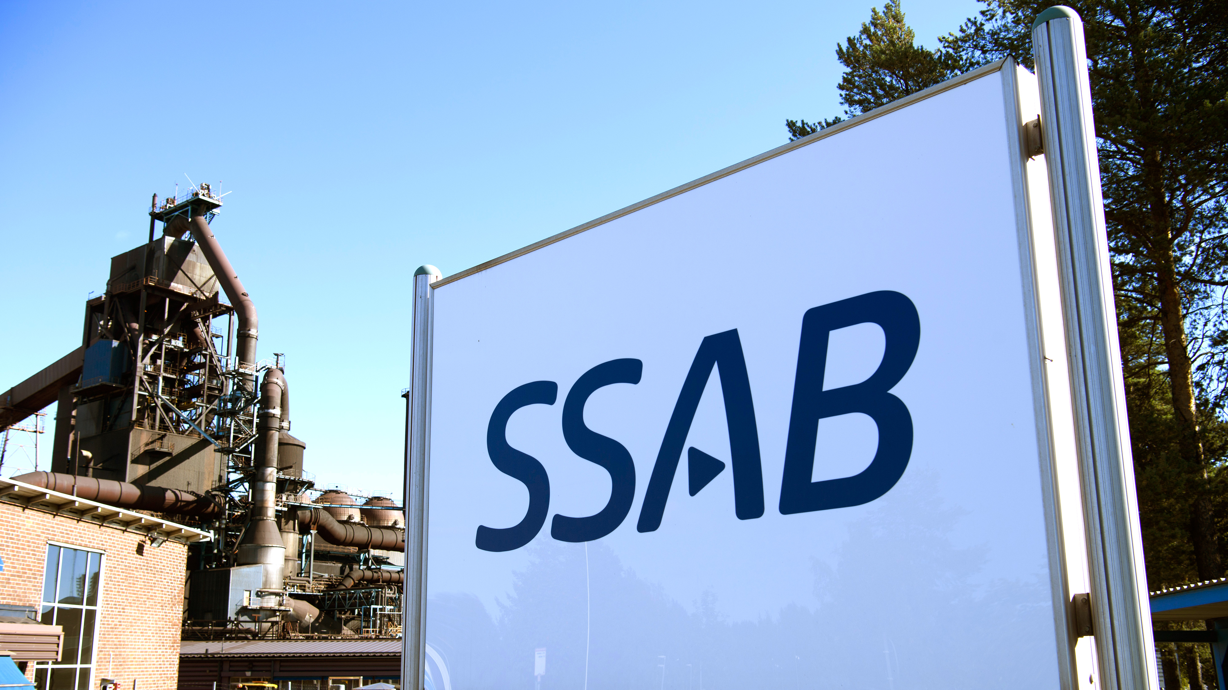 SSAB-skylten står utomhus bredvid en industribyggnad med metallkonstruktioner och träd i bakgrunden.