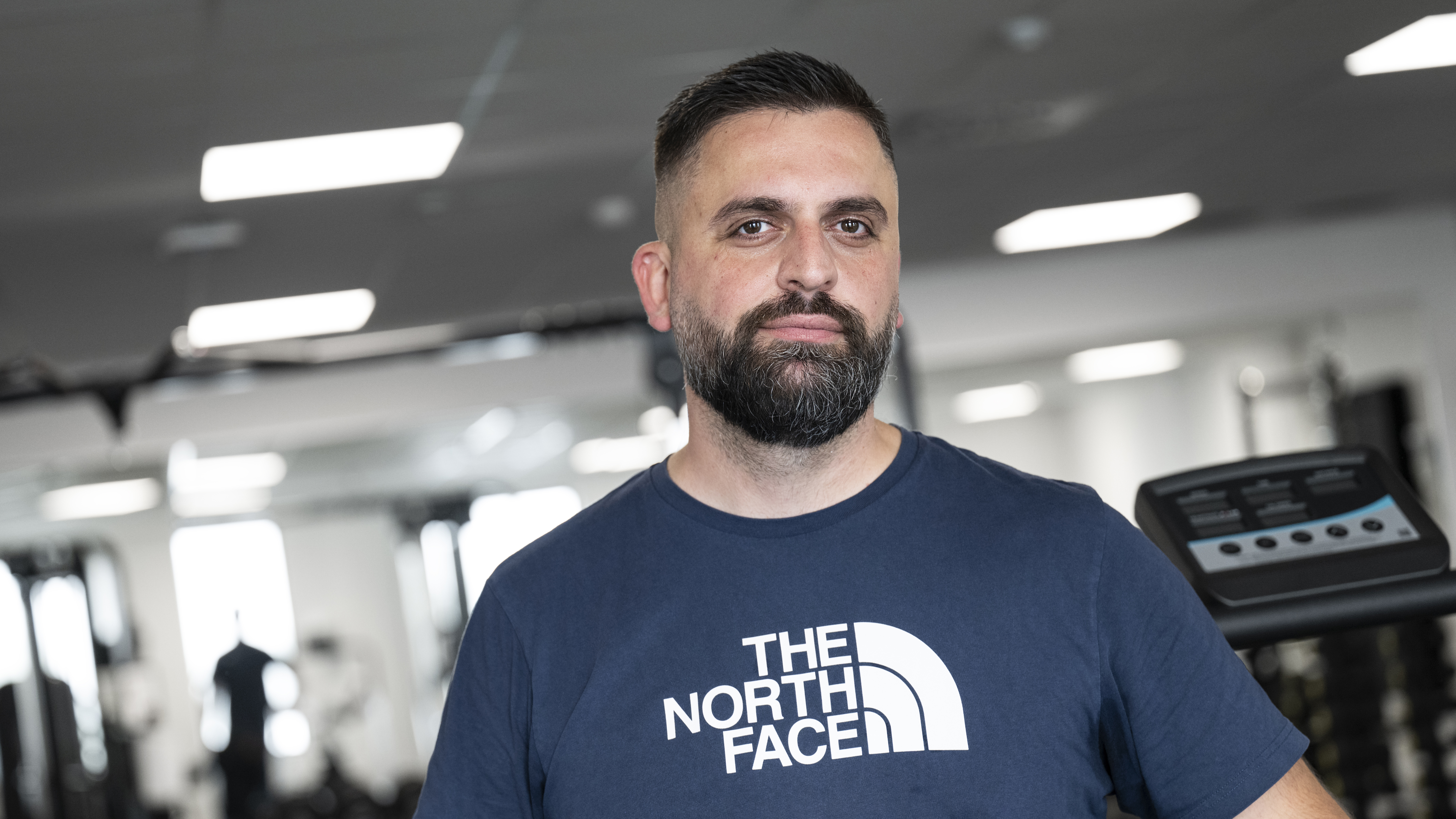 Afrim Mehmeti med skägg och en mörkblå t-shirt står i ett gym där träningsutrustning syns i bakgrunden.
