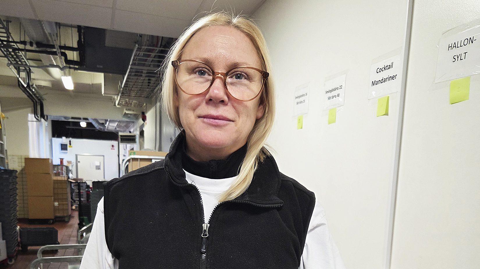 Jessica Hahne med blont hår och stora glasögon står inomhus nära en vägg med etiketter och hyllor i bakgrunden.