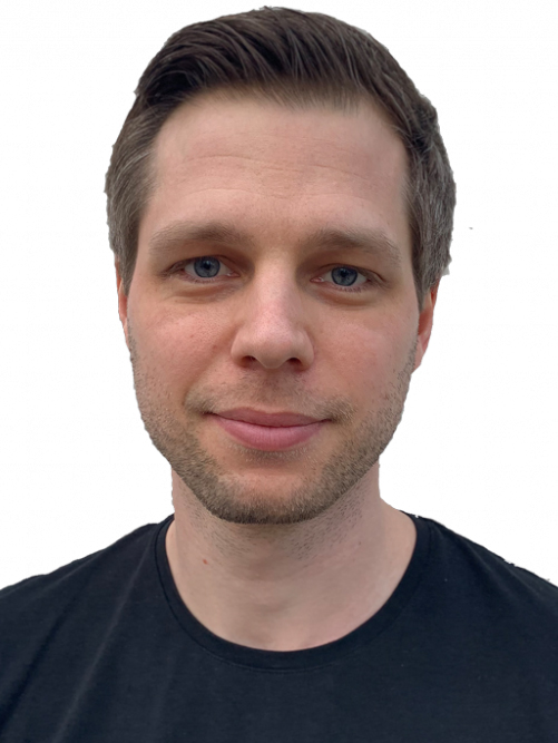 En man med kort brunt hår och ljus hud ser mot kameran. Han har på sig en svart t-shirt och står framför en vit bakgrund.