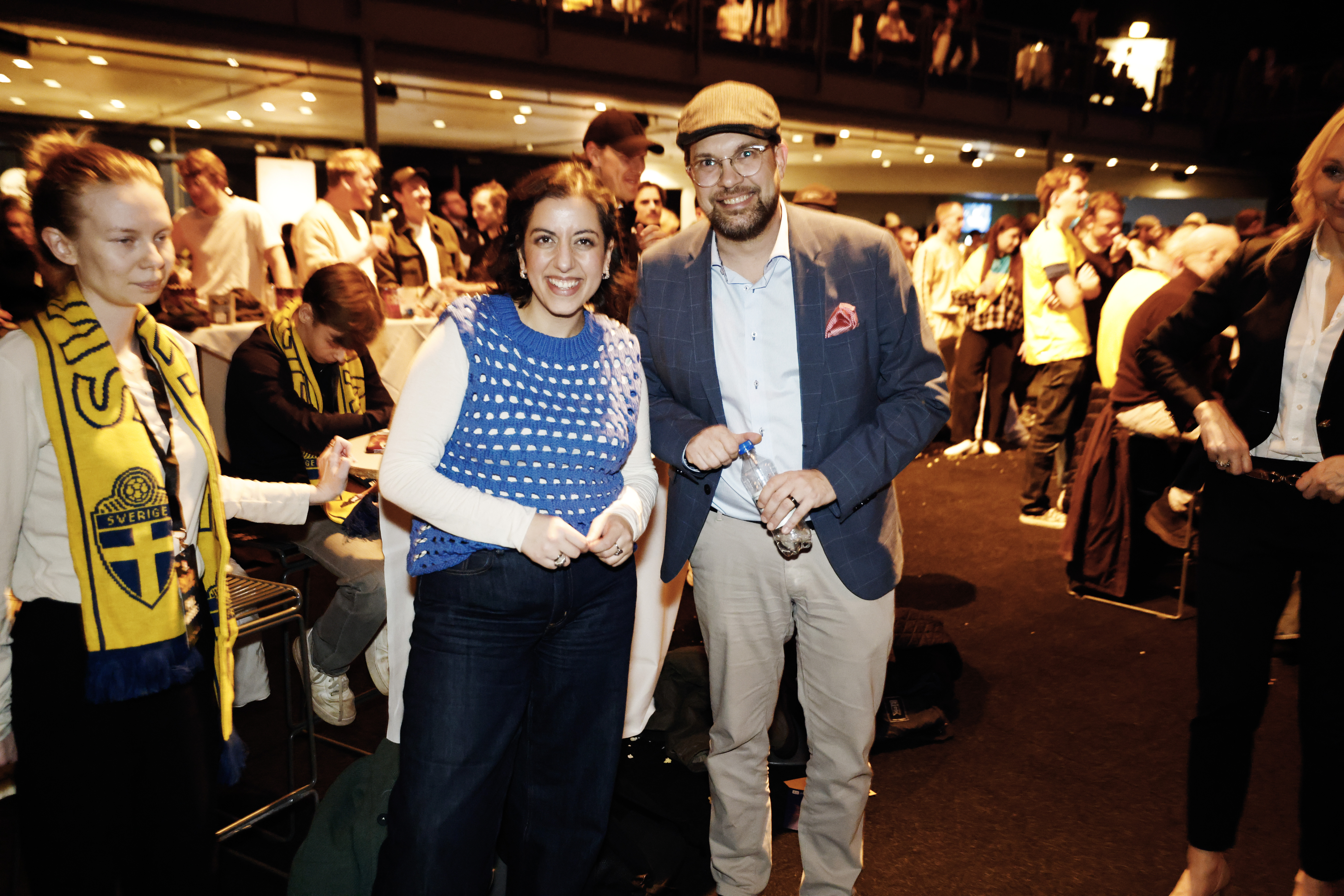 Två personer poserar och ler mot kameran på ett inomhusevenemang. Andra i bakgrunden har gula halsdukar och samtalar.