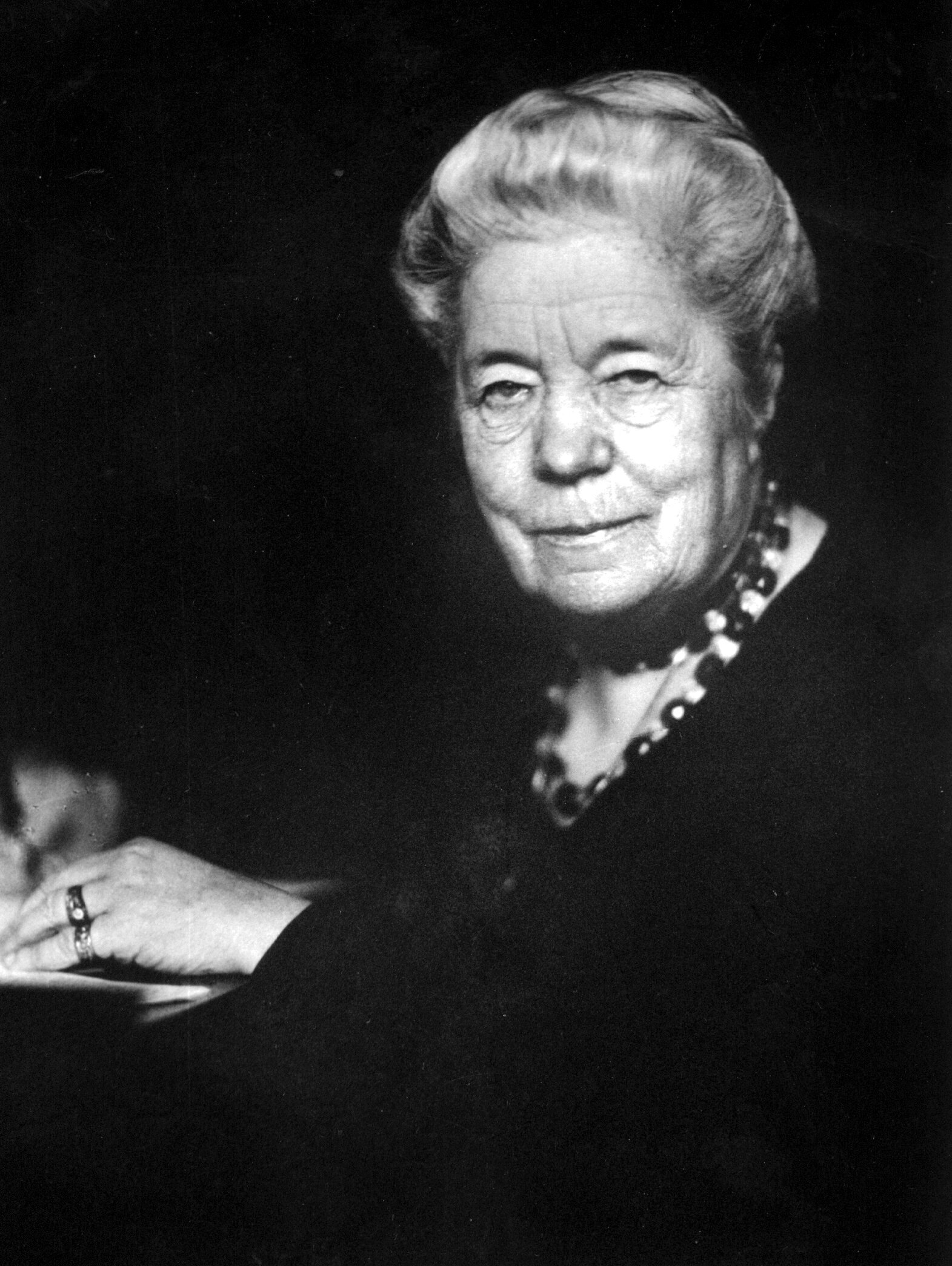 Äldre kvinna med uppsatt ljust hår, bär mörk klänning och pärlhalsband, sitter och ser mot kameran med svag leende.