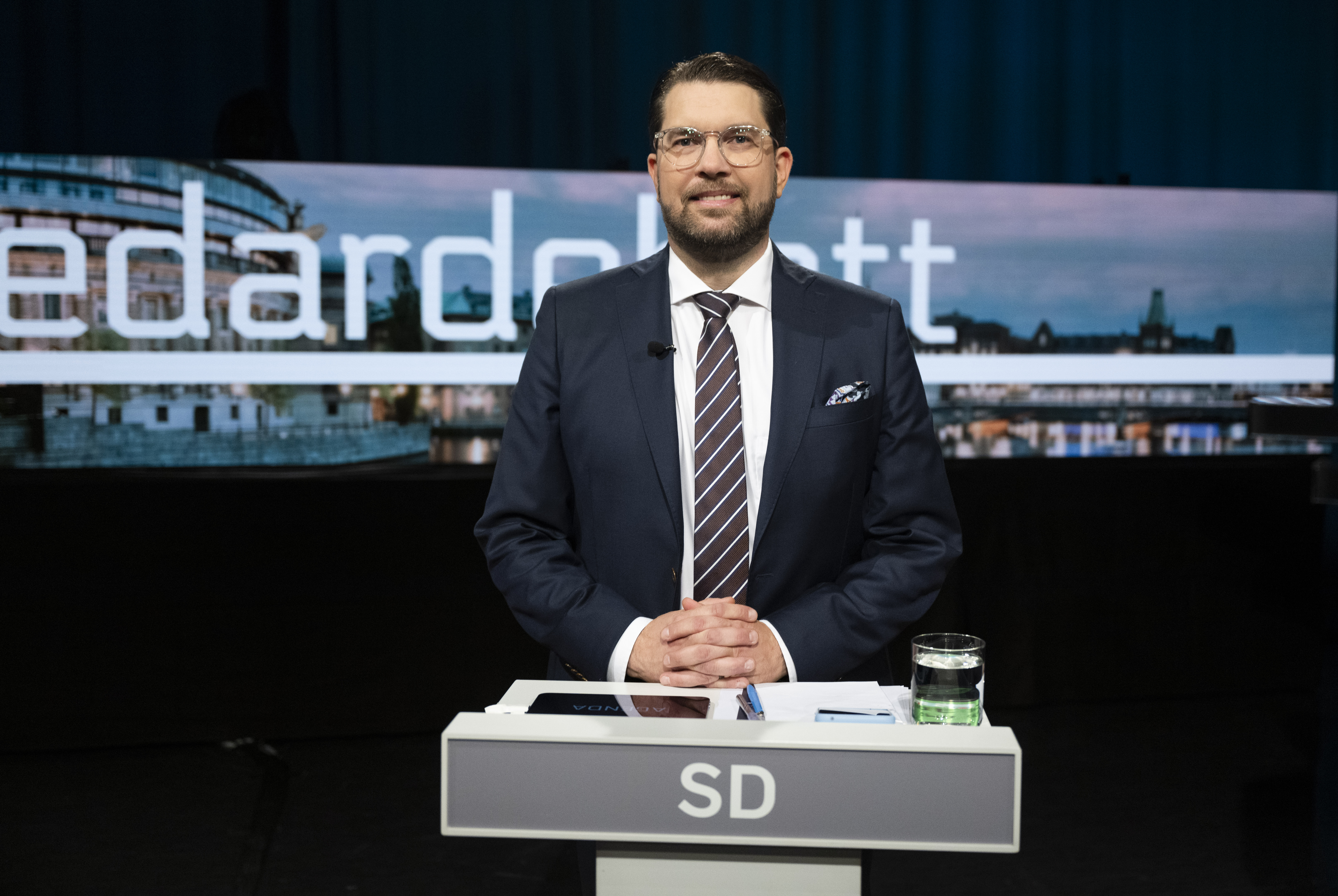 En man i kostym står bakom ett podium märkt "SD" med ett glas vatten, papper och penna. Bakgrunden har en stadssilhuett och stor text.